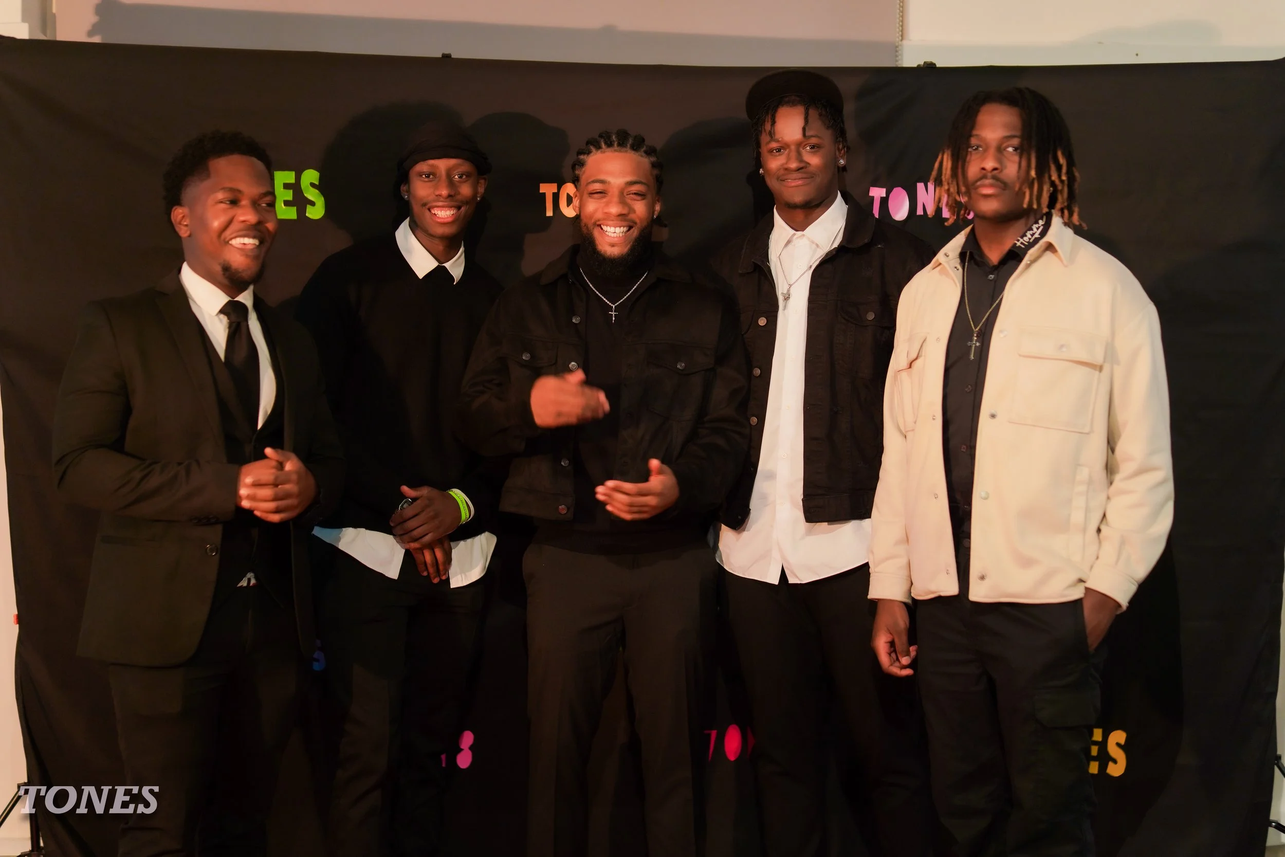 Six young men standing together, dressed in stylish, semi-formal and casual clothes, smiling and posing for a photo indoors against a dark backdrop with colorful text.