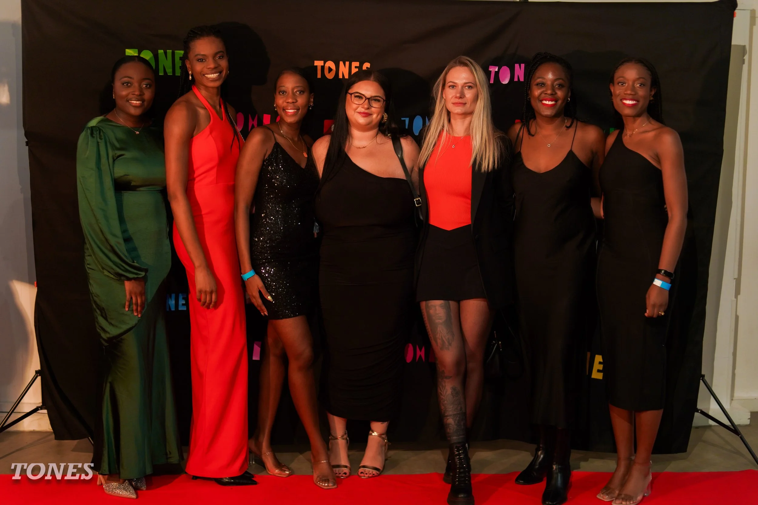 A group of eight women standing together on a red carpet in front of a black backdrop with colorful letters spelling 'TONES'. The women are dressed in elegant evening wear, with some wearing dresses in colors like green, red, black, and gold, and various accessories. They are smiling and posing for the photo.