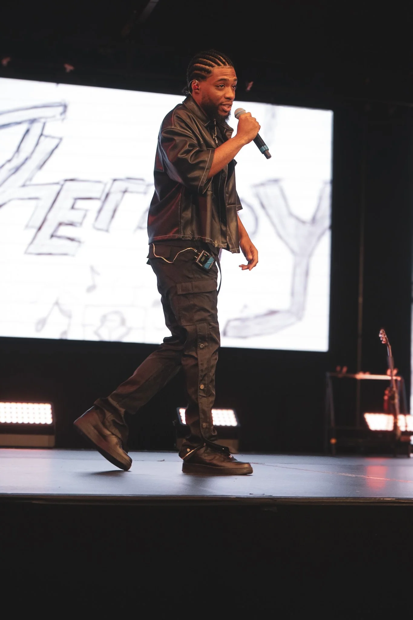 A performer on stage holding a microphone, wearing black cargo pants and a black leather jacket, with a large digital screen behind displaying the word 'HEY'.