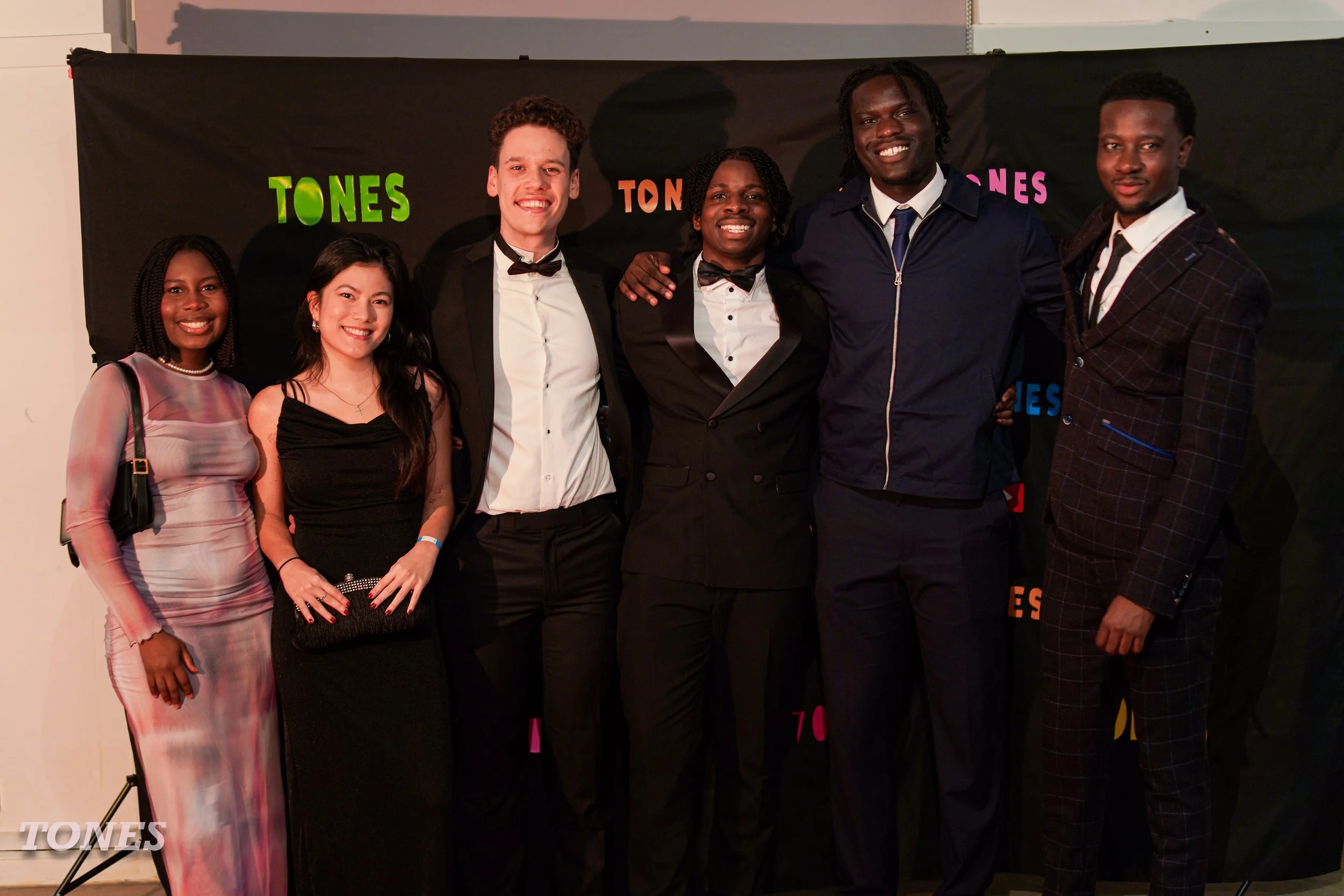 Group of seven diverse young adults dressed in formal attire, standing together and smiling in front of a black backdrop with colorful words, at an event named TONES.