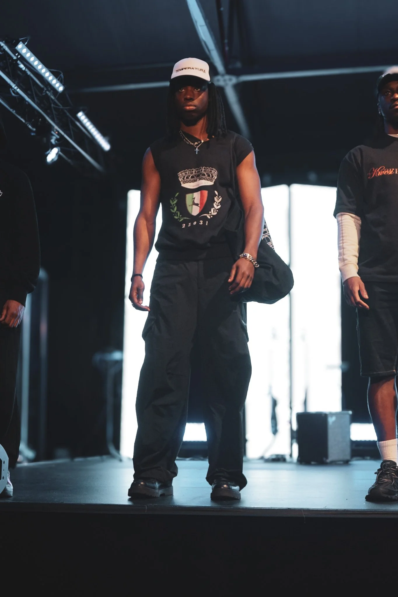 A person standing on a stage wearing a black T-shirt with a crest and the number 25031, black cargo pants, a white cap, and accessories, with two other individuals partially visible beside them.