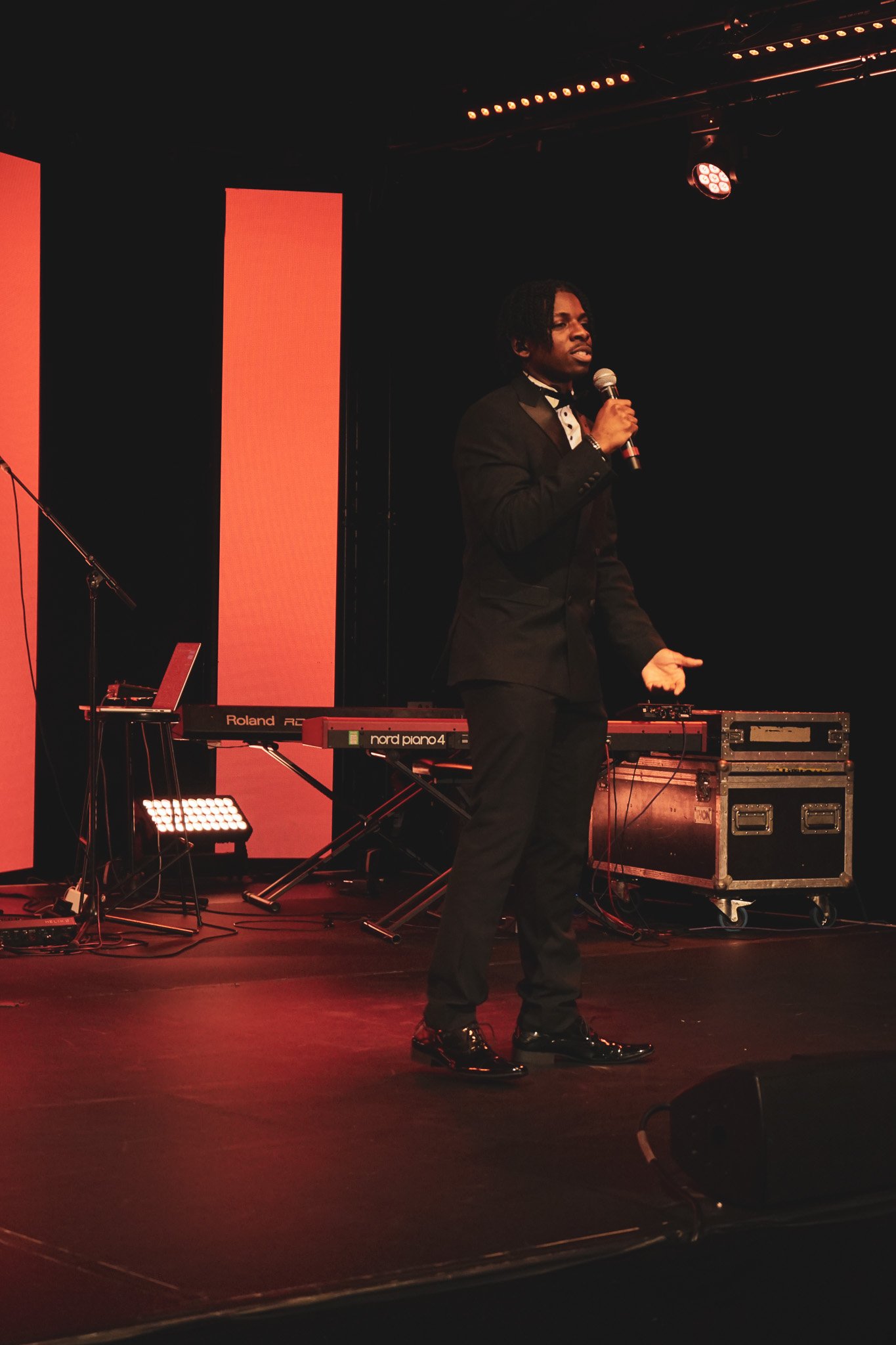A performer dressed in a black tuxedo with a bow tie, holding a microphone and singing on stage, with musical equipment including a keyboard, laptop, and sound equipment behind him.