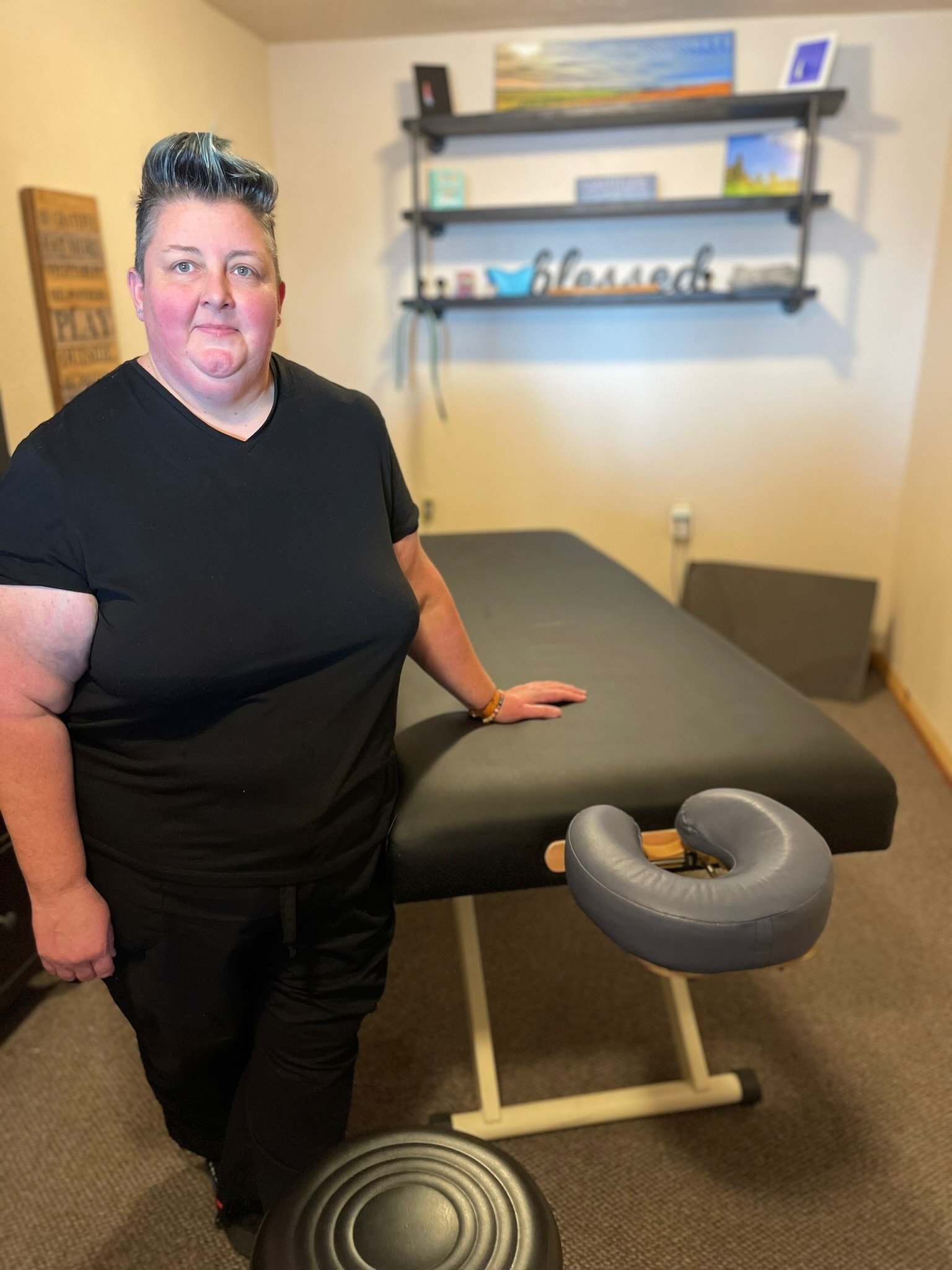 A woman with short hair wearing a black t-shirt and black pants standing in a room with a massage or therapy table, a black stool, and decorative wall shelves with artwork and decor.