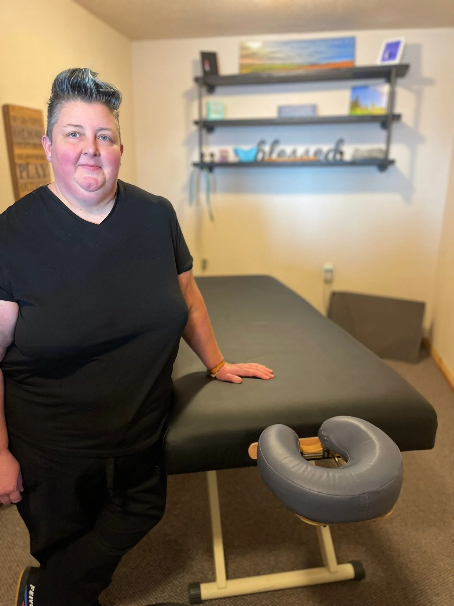 A person with short, styled hair standing next to a massage or therapy table in a room with decorative shelves and wall art.