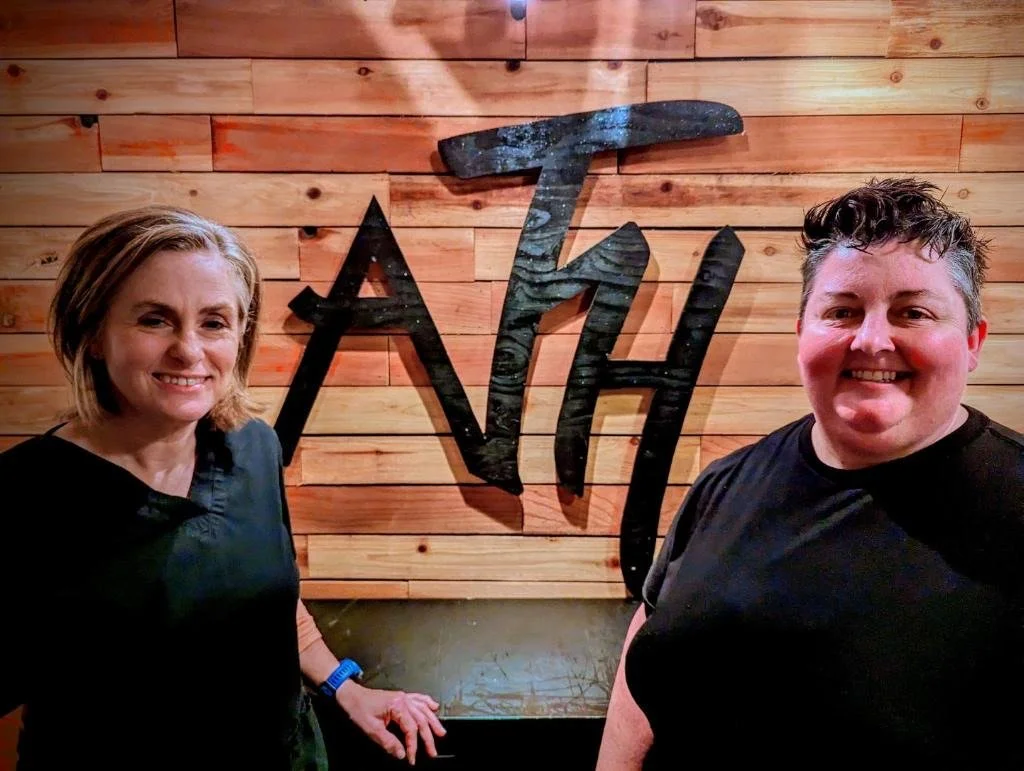 A woman and a man smiling and standing in front of a wooden wall with a large black metal decorative letter display reading 'A7H'.