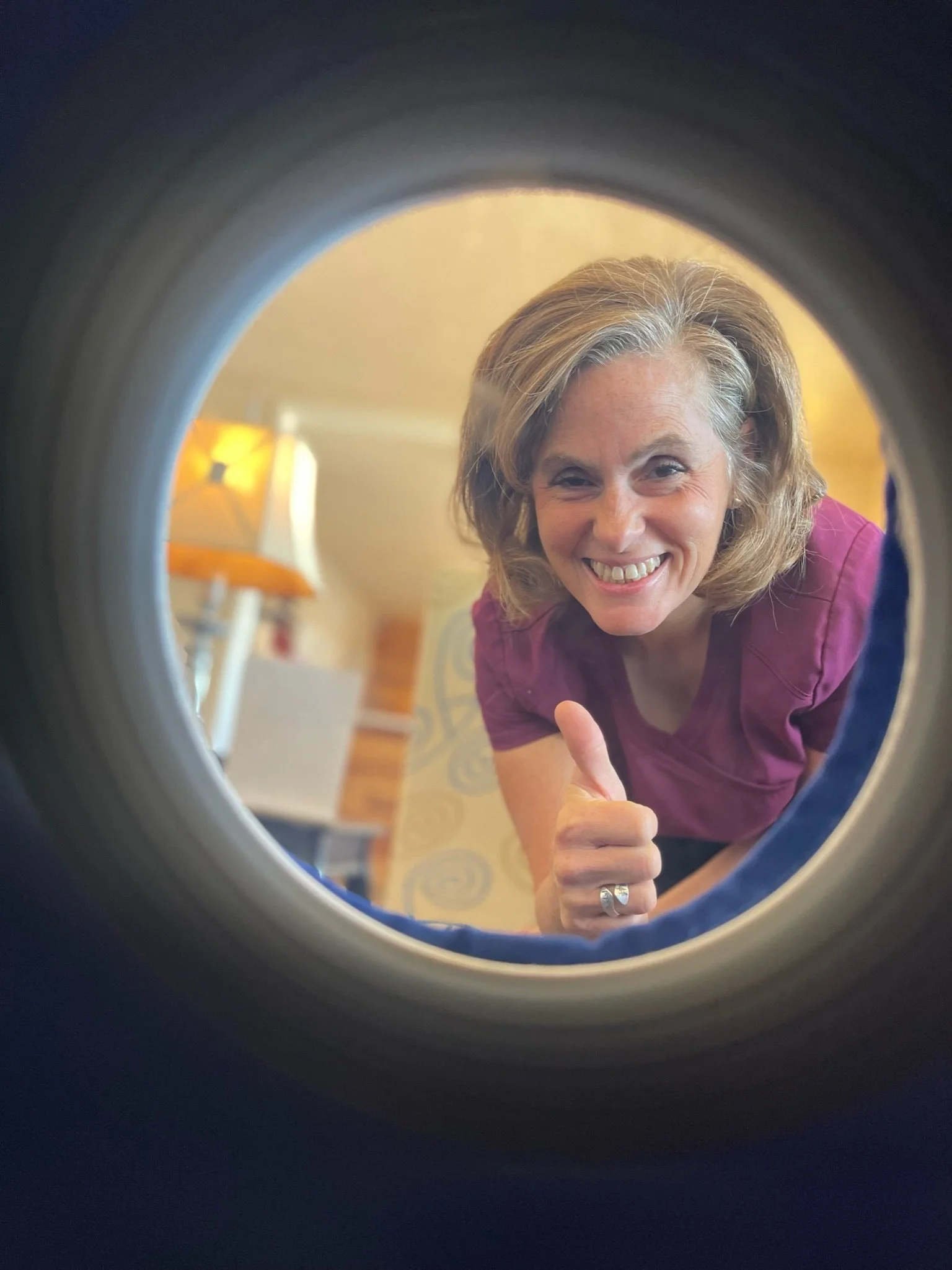 Woman smiling and giving a thumbs-up through a circular opening, with a home interior in the background.