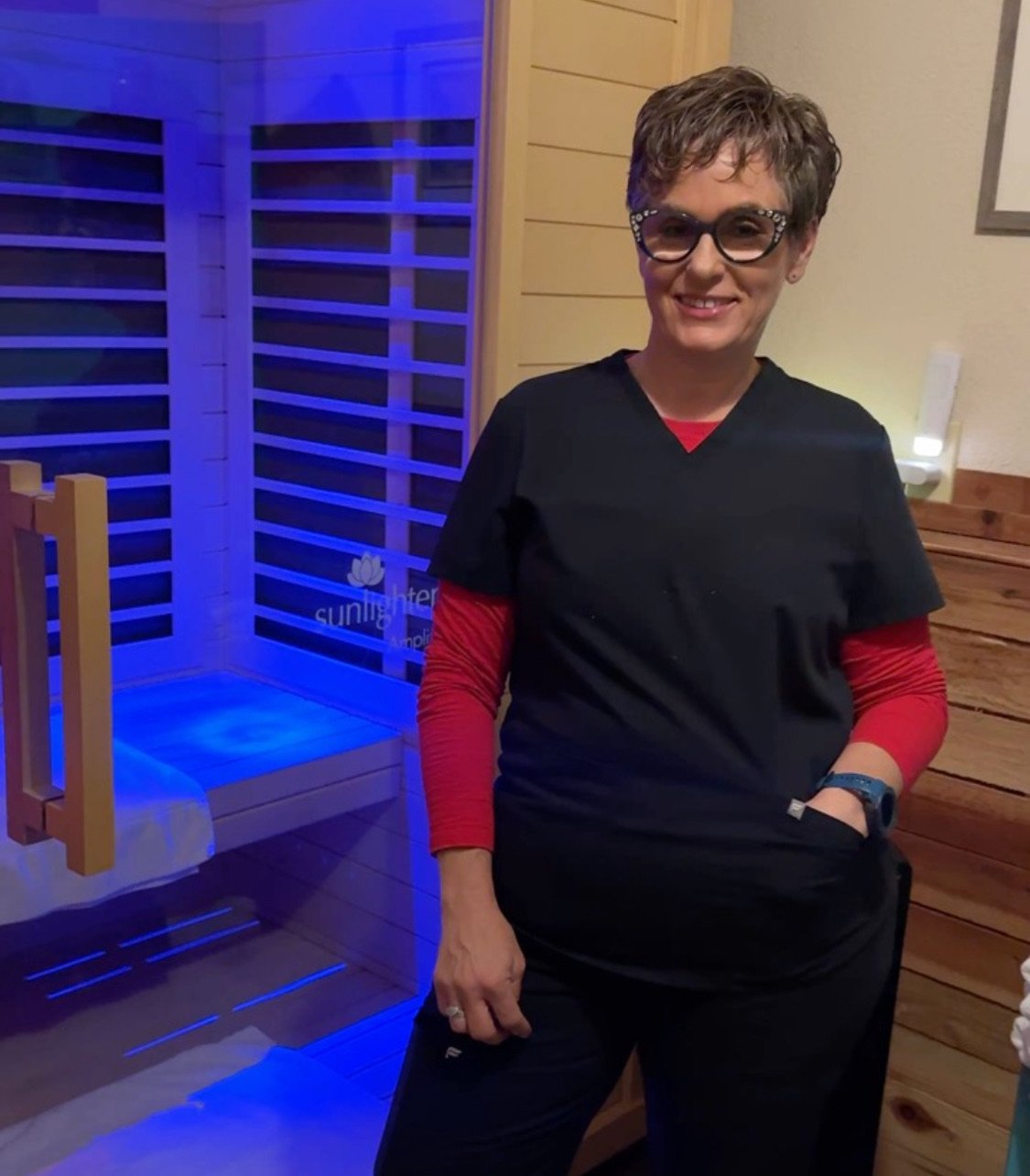 A woman with short hair, glasses, and a black and red outfit is smiling indoors near a wooden wall and a blue-lit sauna with the brand name Sunlight visible.