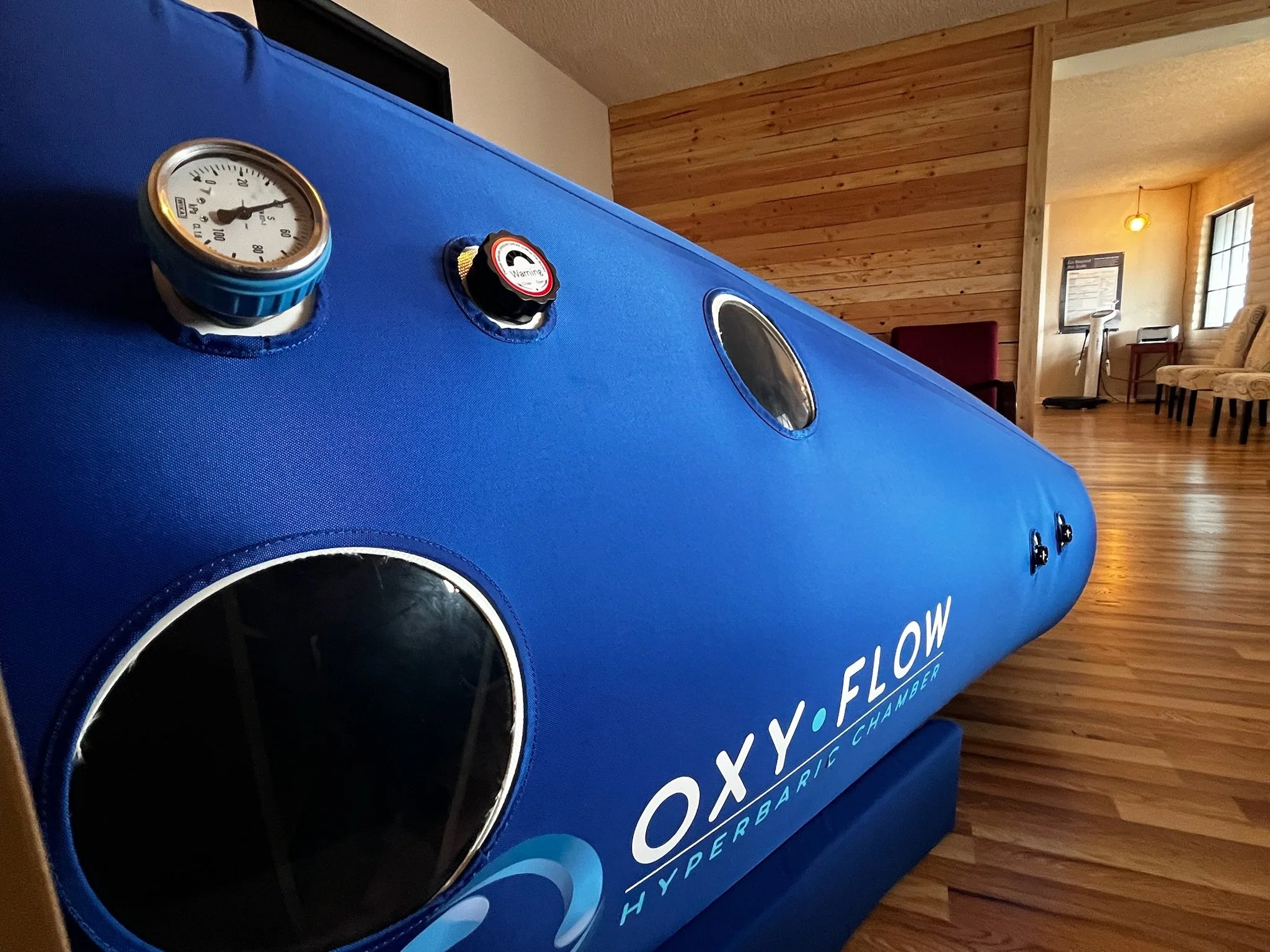 Close-up of a blue hyperbaric oxygen chamber labeled 'OXY.FLOW' with pressure gauge and controls, inside a room with wooden floors and walls, chairs, and large windows in the background.