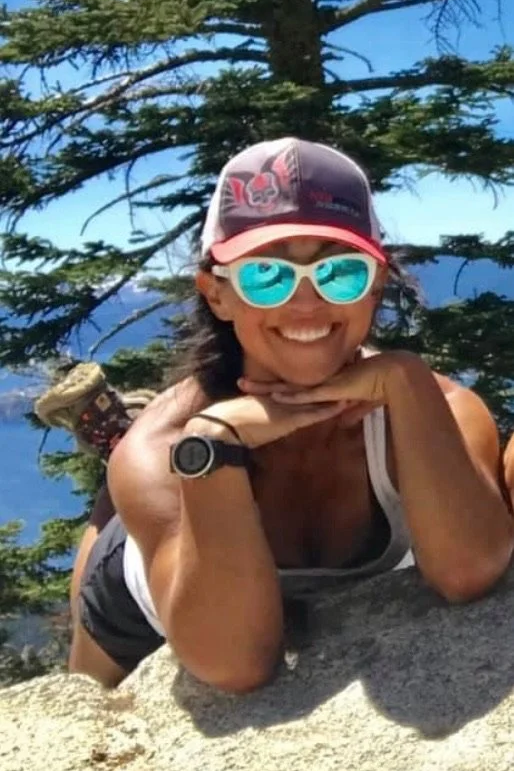 Woman in outdoor setting wearing a cap, sunglasses, and a watch, lying on rocks and smiling at camera, with trees and sky in background.
