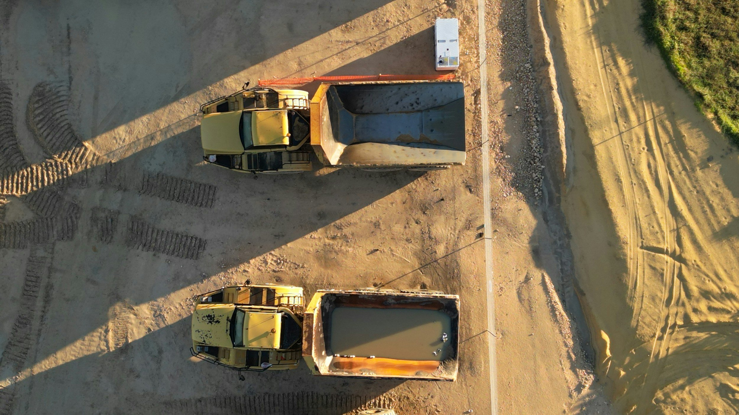 Two yellow dump trucks filled with muddy water are parked on a construction site, with tire tracks visible on the dirt ground and a small white trailer nearby.