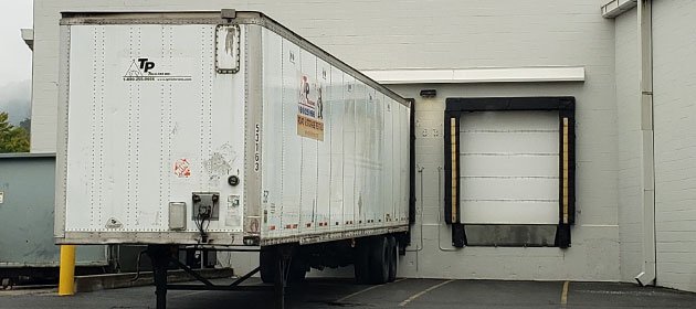 A white semi-truck trailer parked against a building with a white loading dock door.