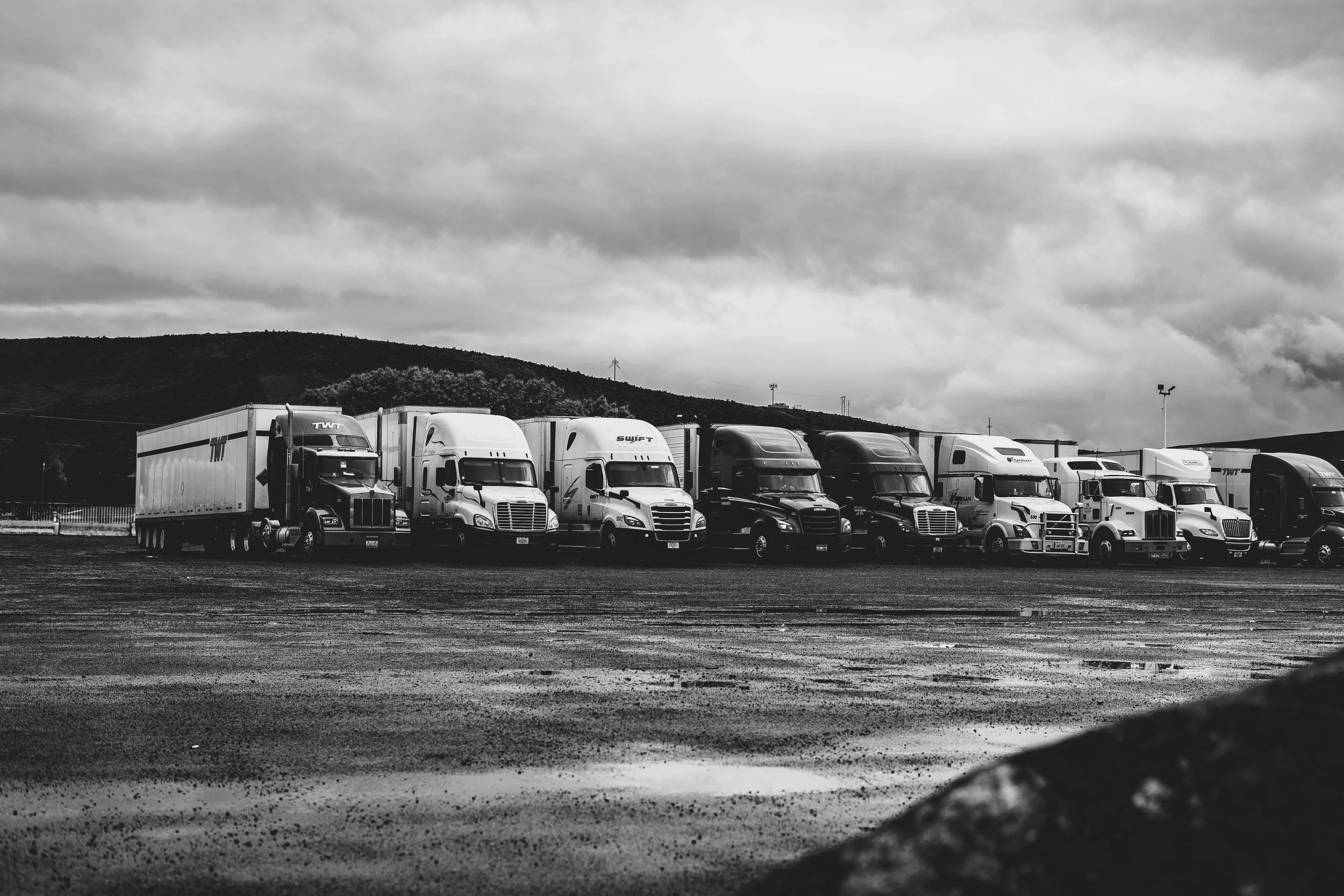 Line of semi-trucks parked in an open lot on a cloudy day, with hills and overcast sky in the background, in black and white.