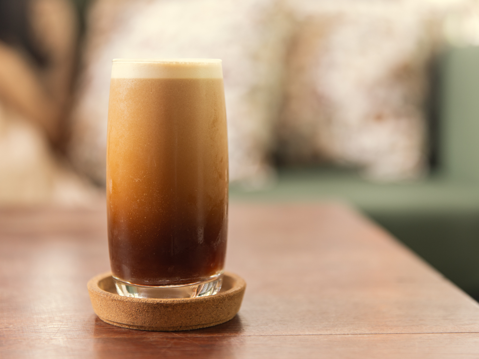 Un verre tallé avec une boisson glacée, probablement un café色 iced咖啡, sur une table en bois avec un sous-verre en liège, dans un environnement intérieur avec un fond flou.