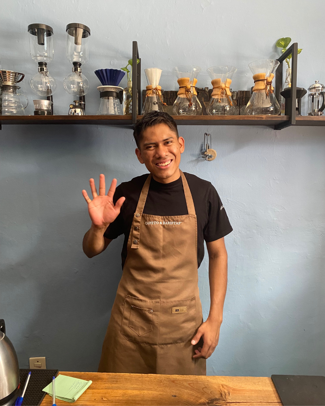 Un homme souriant portant un tablier beige et un t-shirt noir, saluant de la main, dans une cuisine ou un café