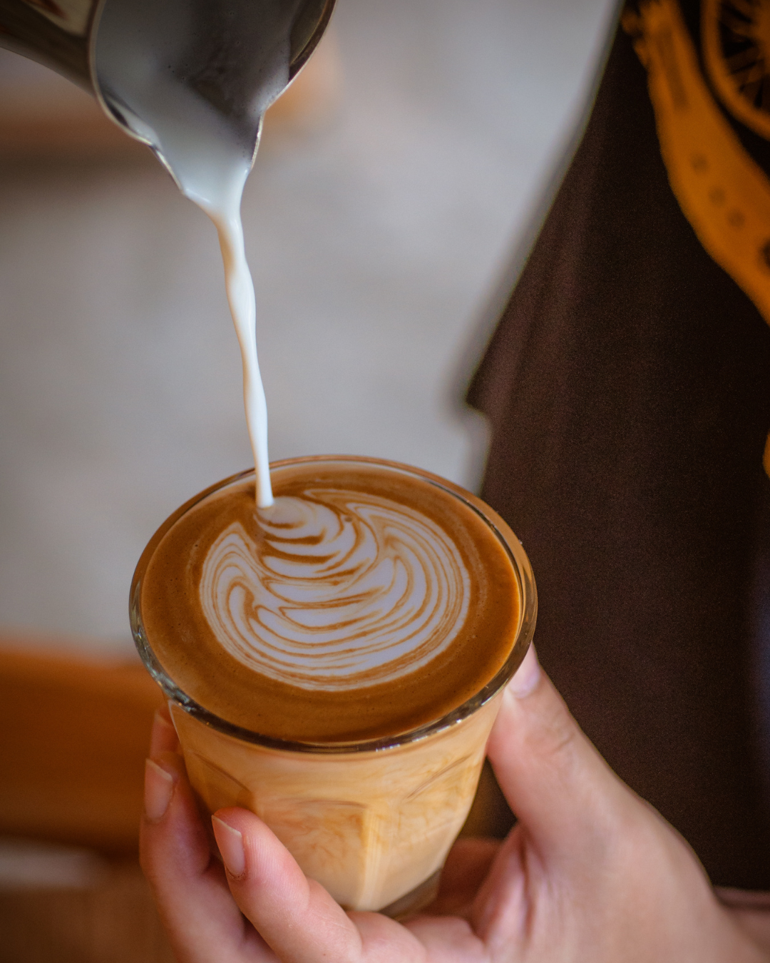 Un barista verse de la crème dans un verre de café latte avec un motif de latte art. La personne tient le verre avec une main et le lait est versé à partir d'une carafe.