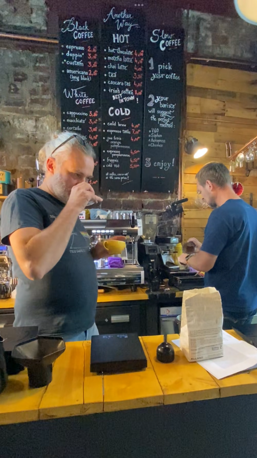 Un comptoir de café avec deux baristas. Un homme teste du café dans une tasse jaune. L'autre homme est en train de préparer un café de spécialoyé. À l'arrière-plan, un panneau noir avec le menu en français et en anglais.
