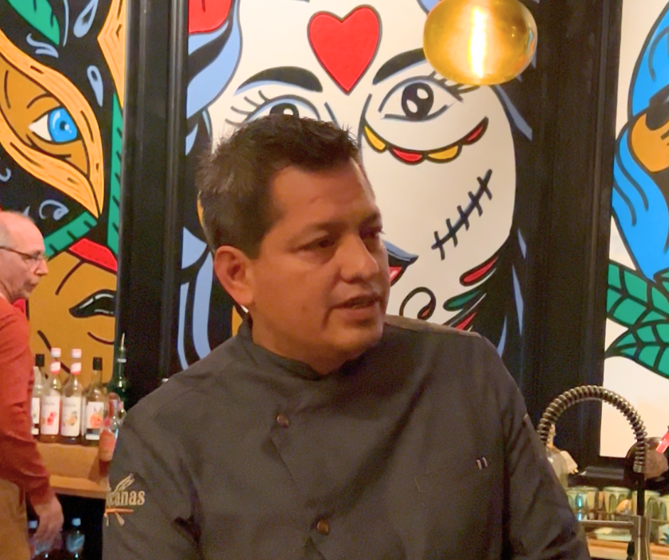 Un homme vêtu d'une veste de chef dans un restaurant avec un mur décoré d'une fresque colorée d'une face de style mexicain contemporain, avec des éléments de dessin cartoon et un visage de femme avec un cœur rouge sur le front.