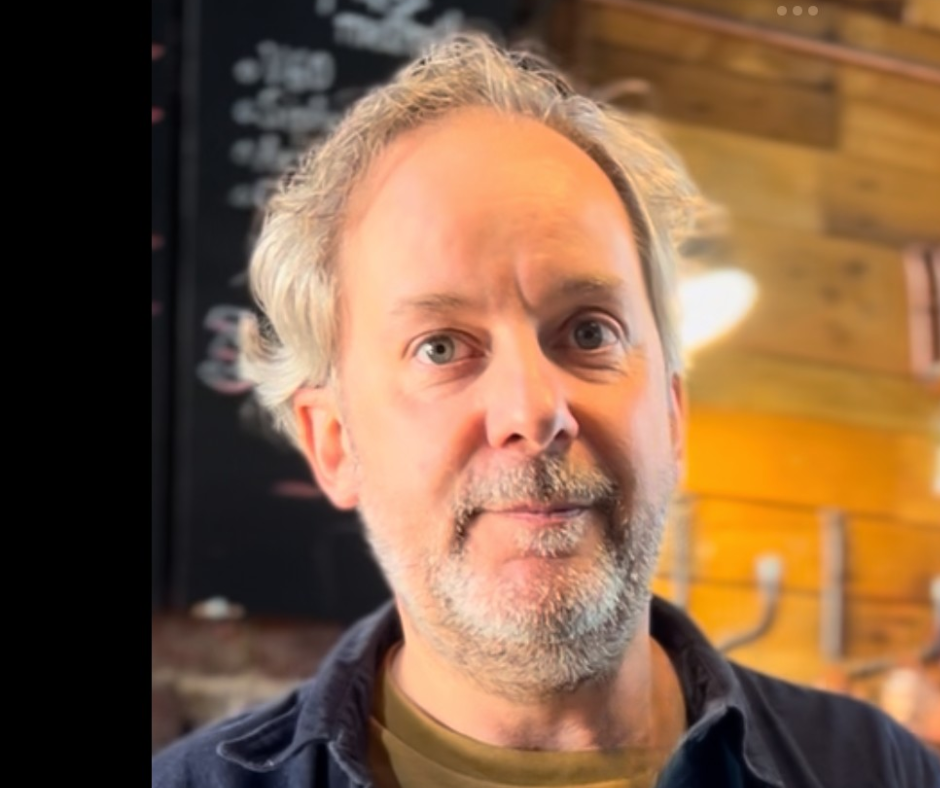 Un homme aux cheveux gris et à la barbe, portant un vêtement foncé, pose pour une photo à l'intérieur d'un établissement, avec un tableau noir dans le fond.