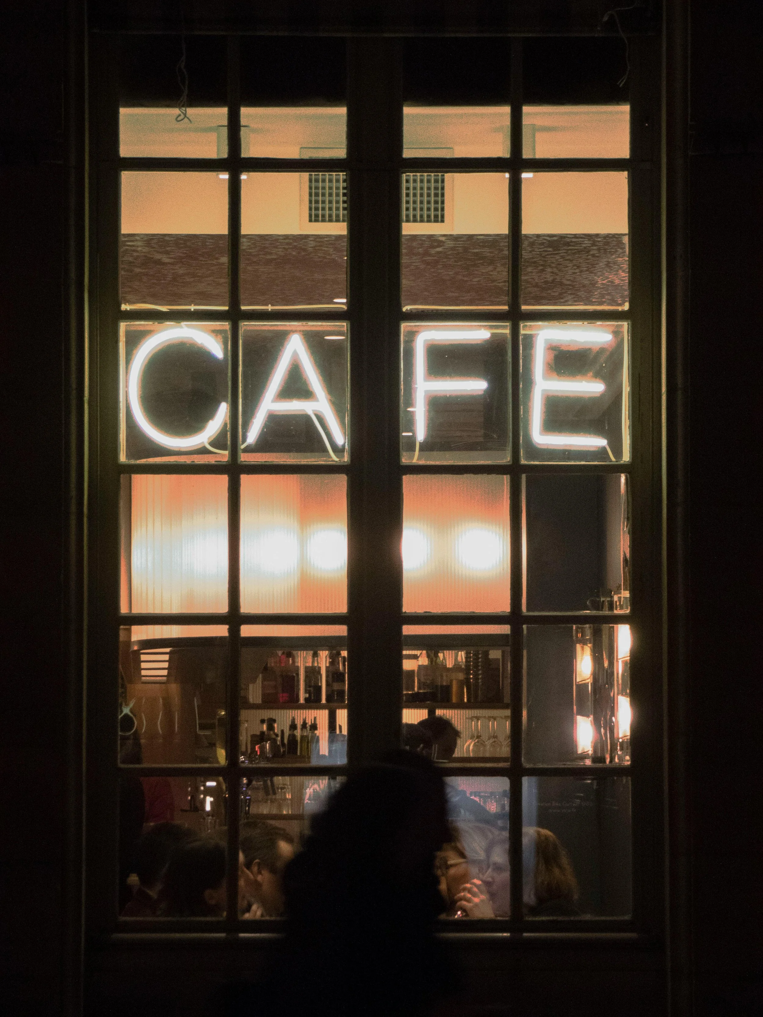 Une fenêtre d'un café avec un panneau lumineux indiquant 'CAFE'. À l'intérieur, des personnes discutent autour de tables, et le bar est visible en arrière-plan.