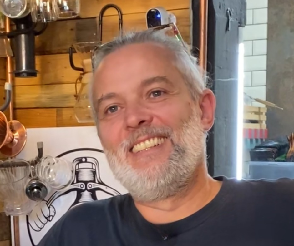 Un homme souriant avec une barbe grise à l'intérieur d'un café, avec des ustensiles de cuisine en cuivre et un dessin stylisé d'une lampe noire sur le mur en bois en arrière-plan.