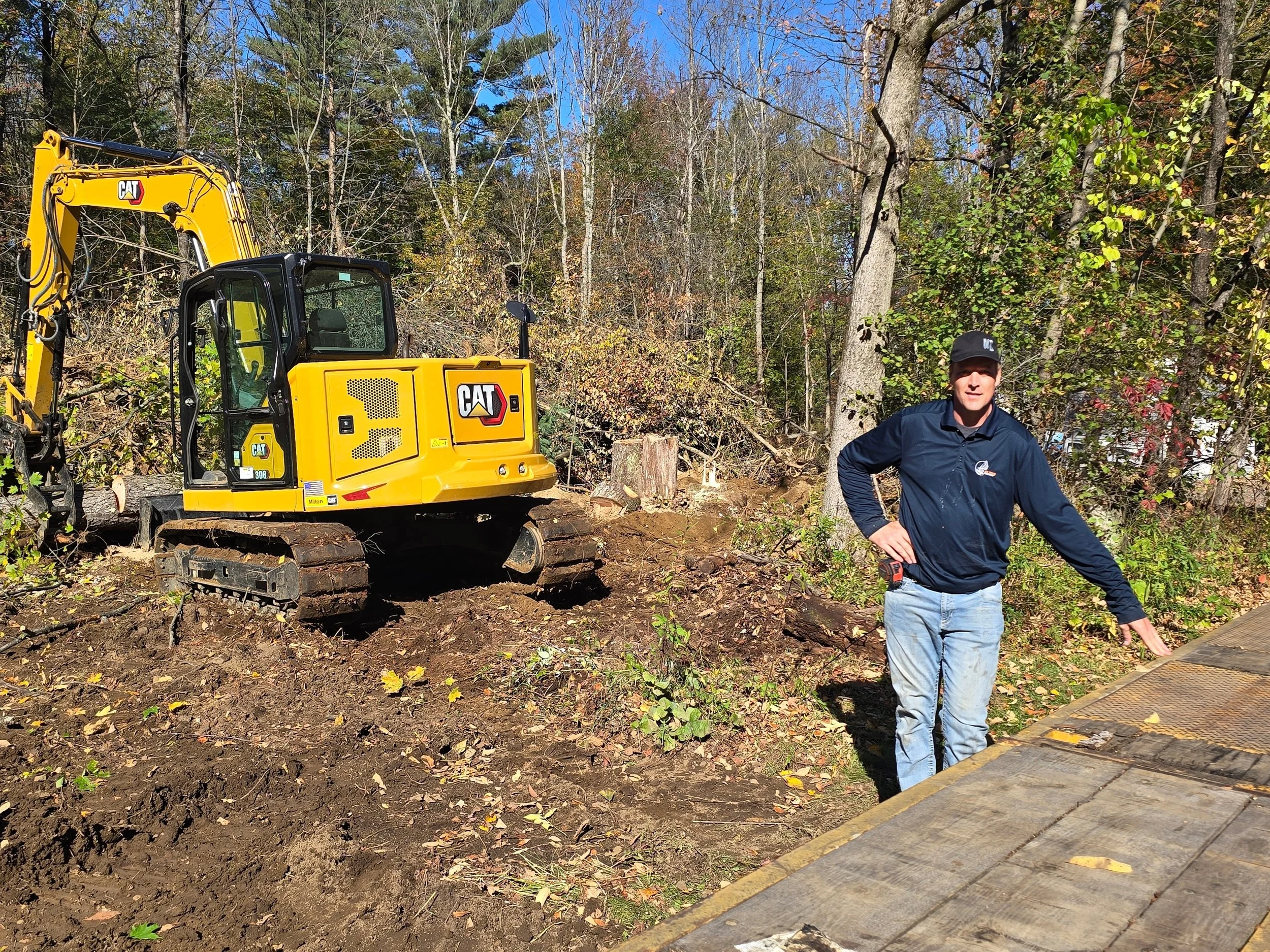 A man wearing a navy jacket and jeans standing outdoors in a wooded area, near a small yellow CAT excavator on a dirt terrain with trees and vegetation in the background.