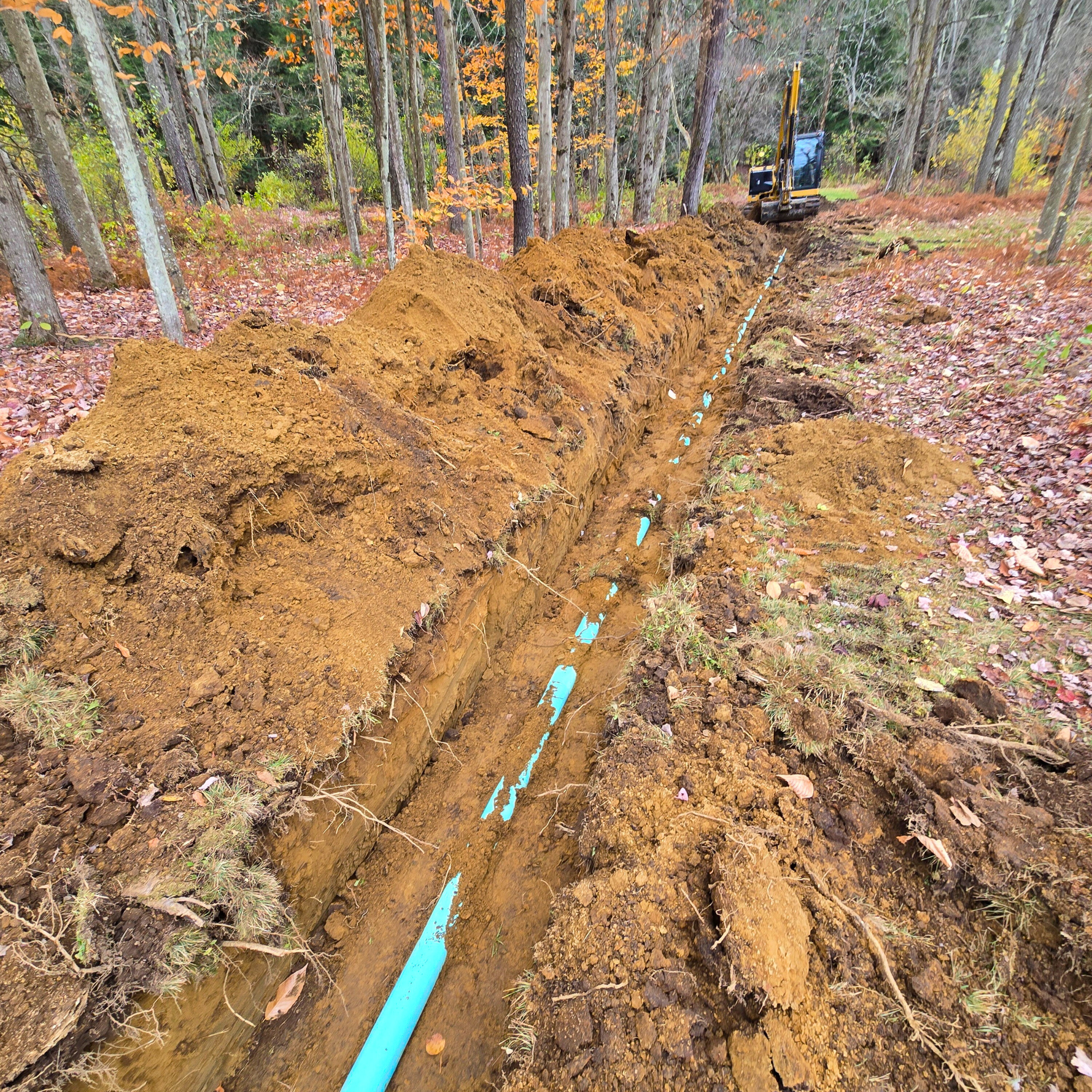 A construction site in a wooded area where a trench has been dug along the length of a blue pipe, with an excavator in the background.