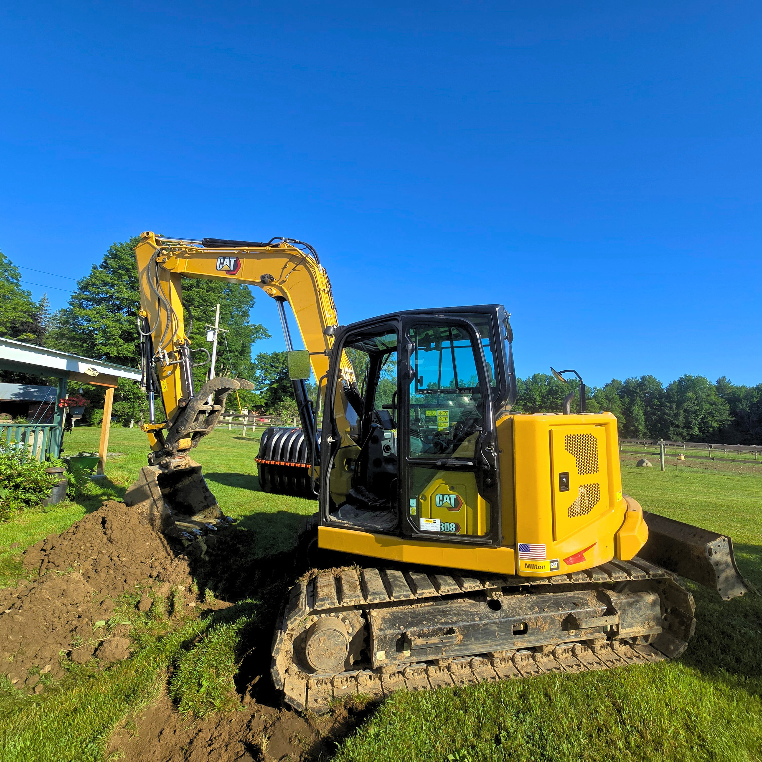 A yellow Caterpillar mini excavator digging into the grass in a backyard, with trees and a clear blue sky in the background.