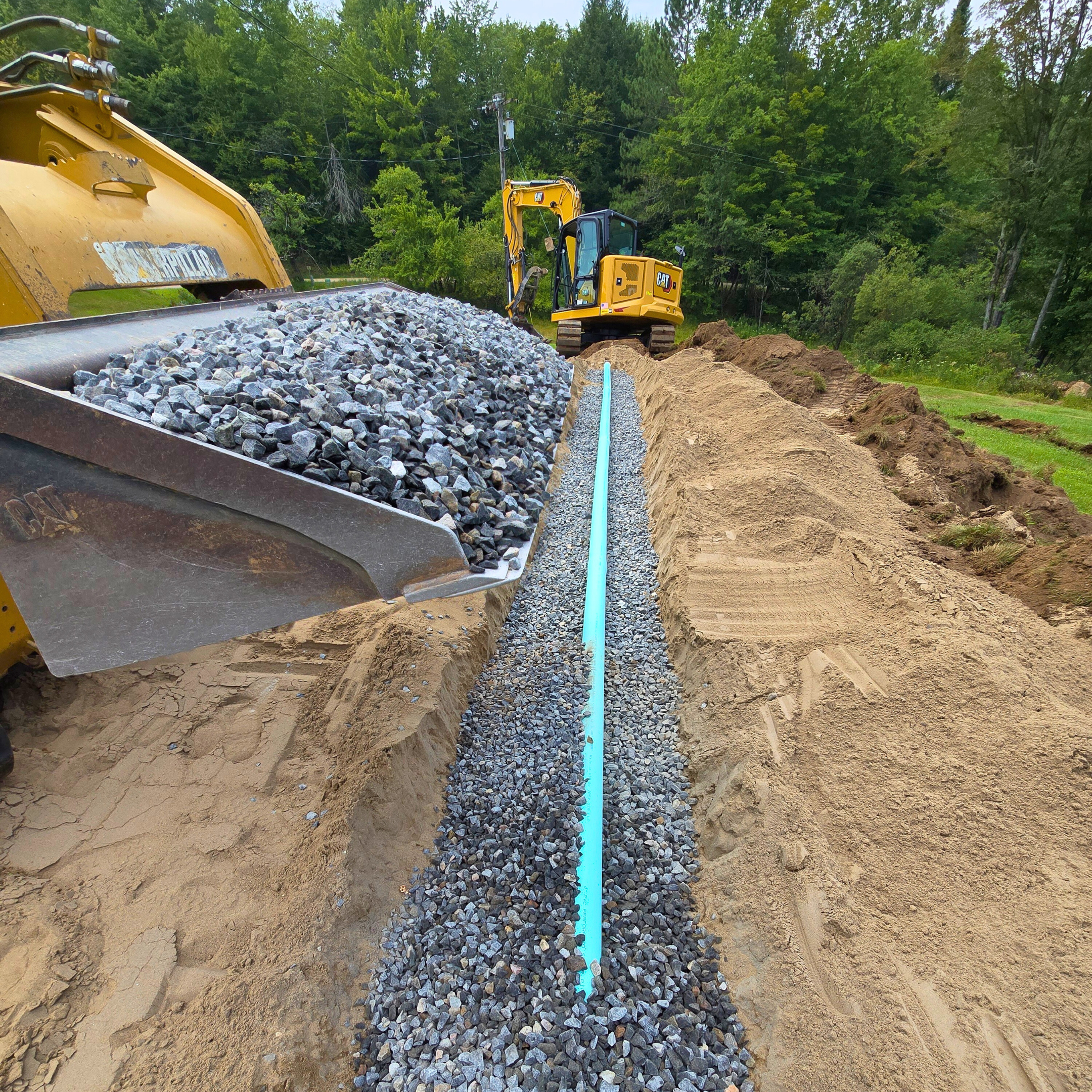 Construction site with a yellow excavator on a dirt mound and a blue pipe installed in a gravel trench surrounded by sand and soil, with trees and power lines in the background.