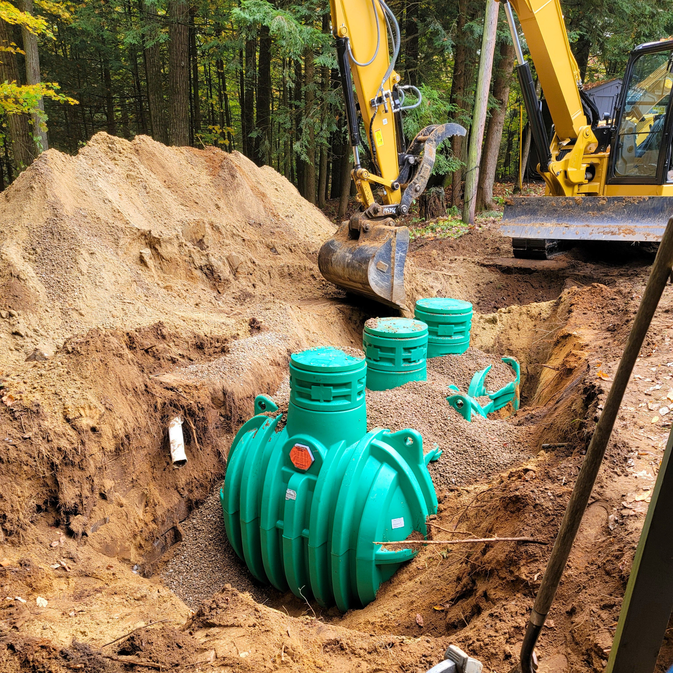 Installation of underground piping with large green fittings and an excavator in a forested area.