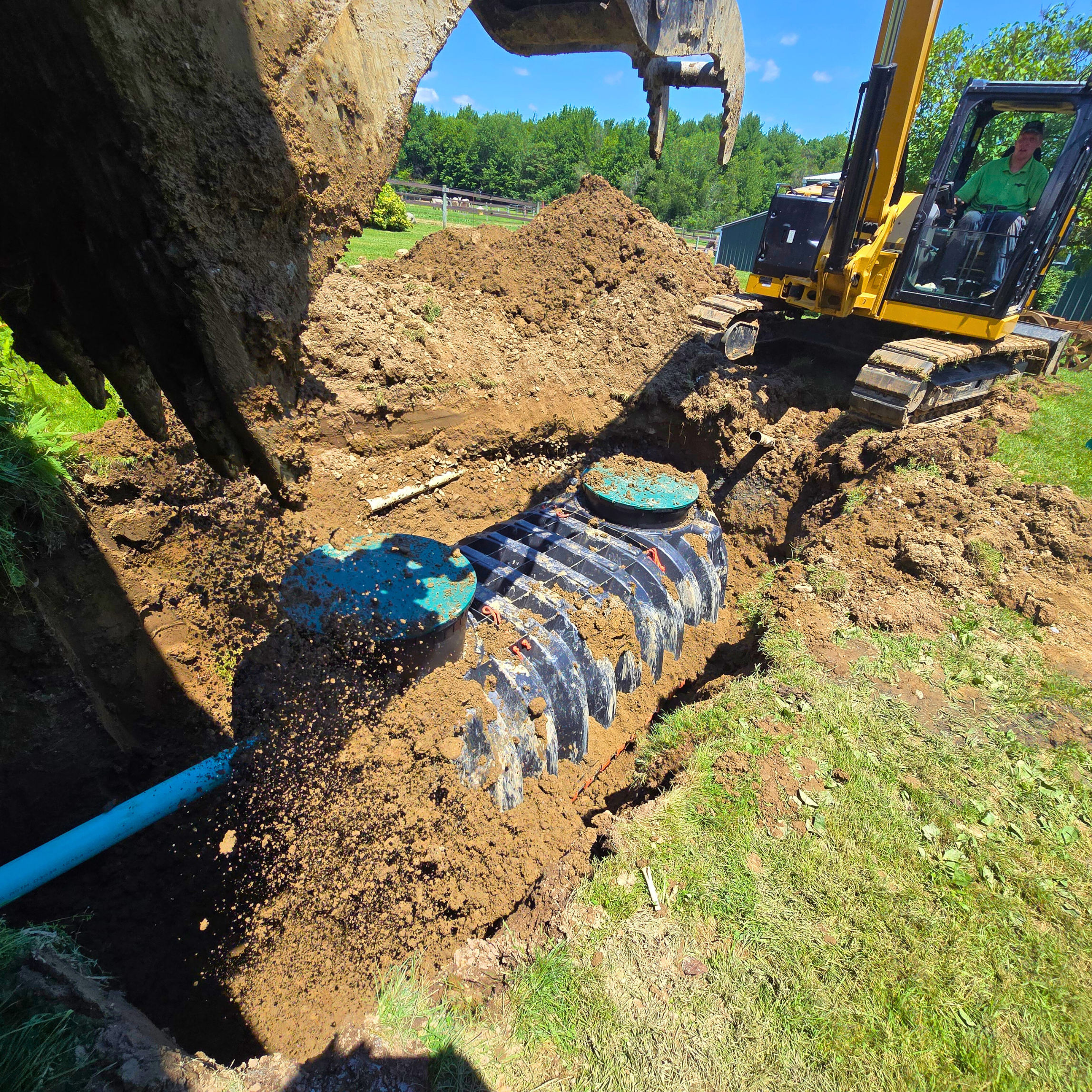 A yellow excavator digging a trench in the ground and installing a large underground septic tank system with black tanks and blue lids in a grassy area on a bright, sunny day.