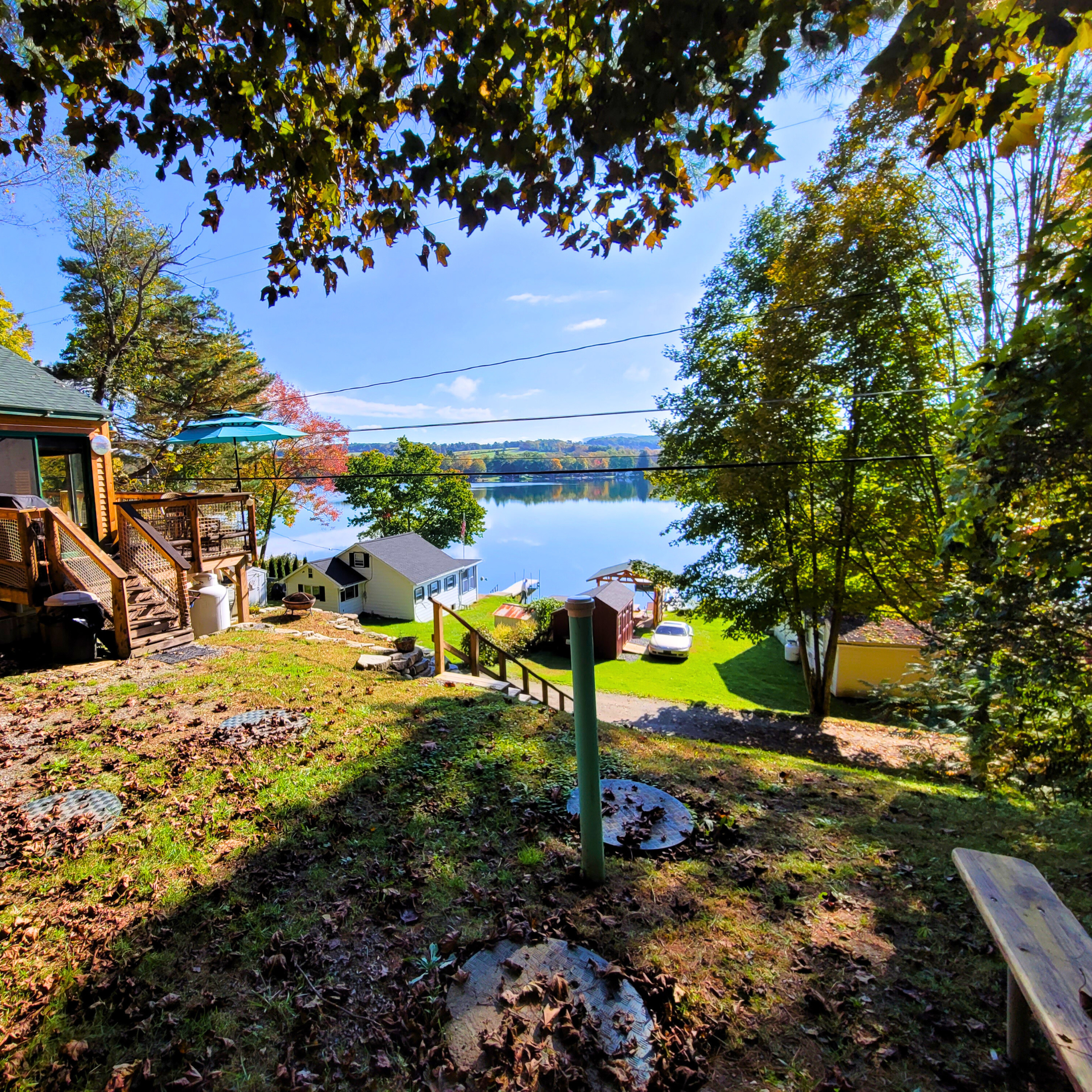 View of a lakeside backyard with trees, a deck, small houses, a dock, a car, and a clear blue sky reflected in the water.