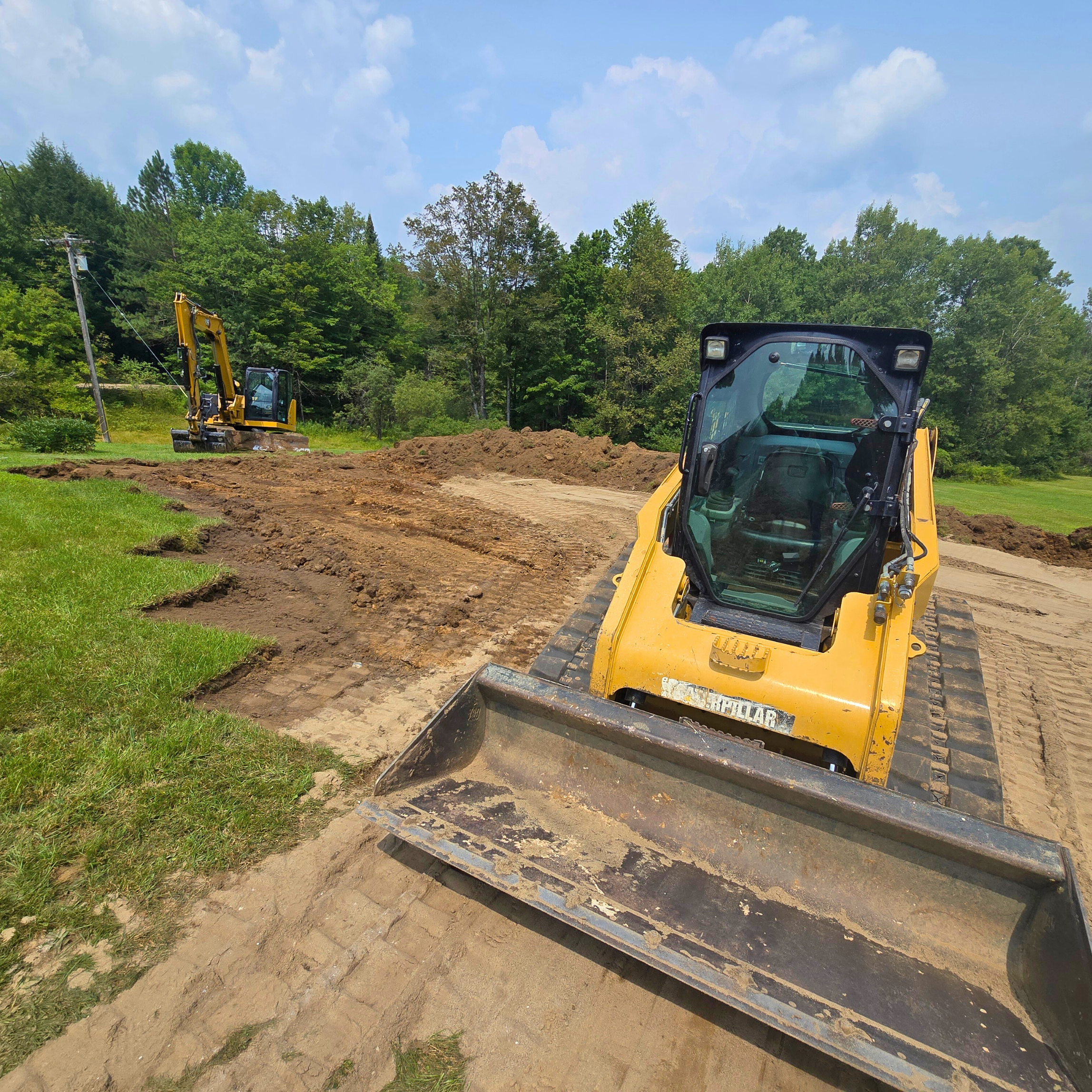 Construction site with two yellow bulldozers leveling ground on a partially grassy area, surrounded by trees under a partly cloudy sky.