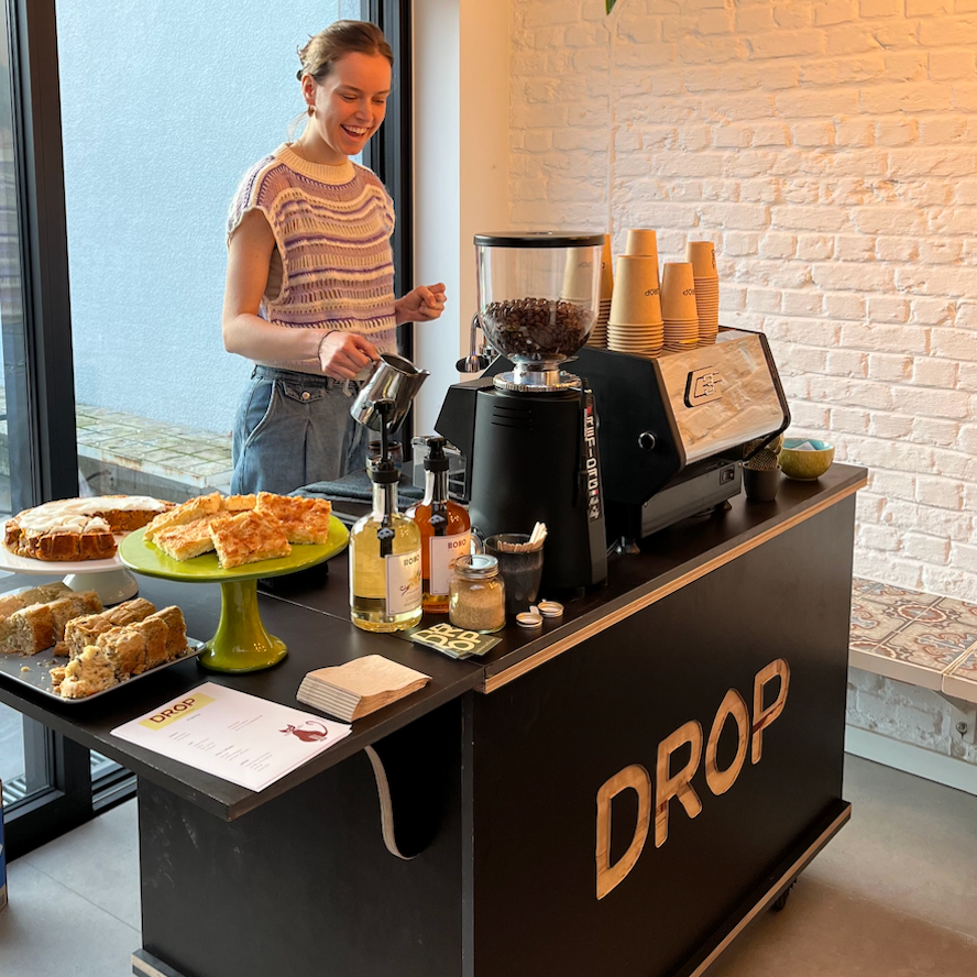 A barista preparing coffee at a mobile coffee cart station with baked goods, bottles of syrup, cups, and a menu on the counter.