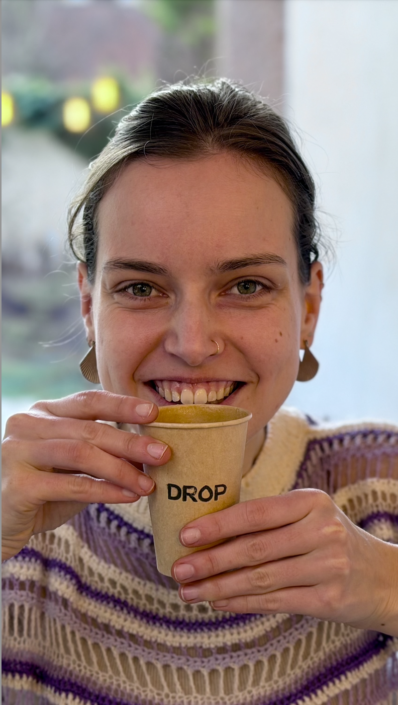 The owner and barista of the mobile coffee cart Drop. She is holding an oat flat white in a paper coffee cup, with the name of her company on it.