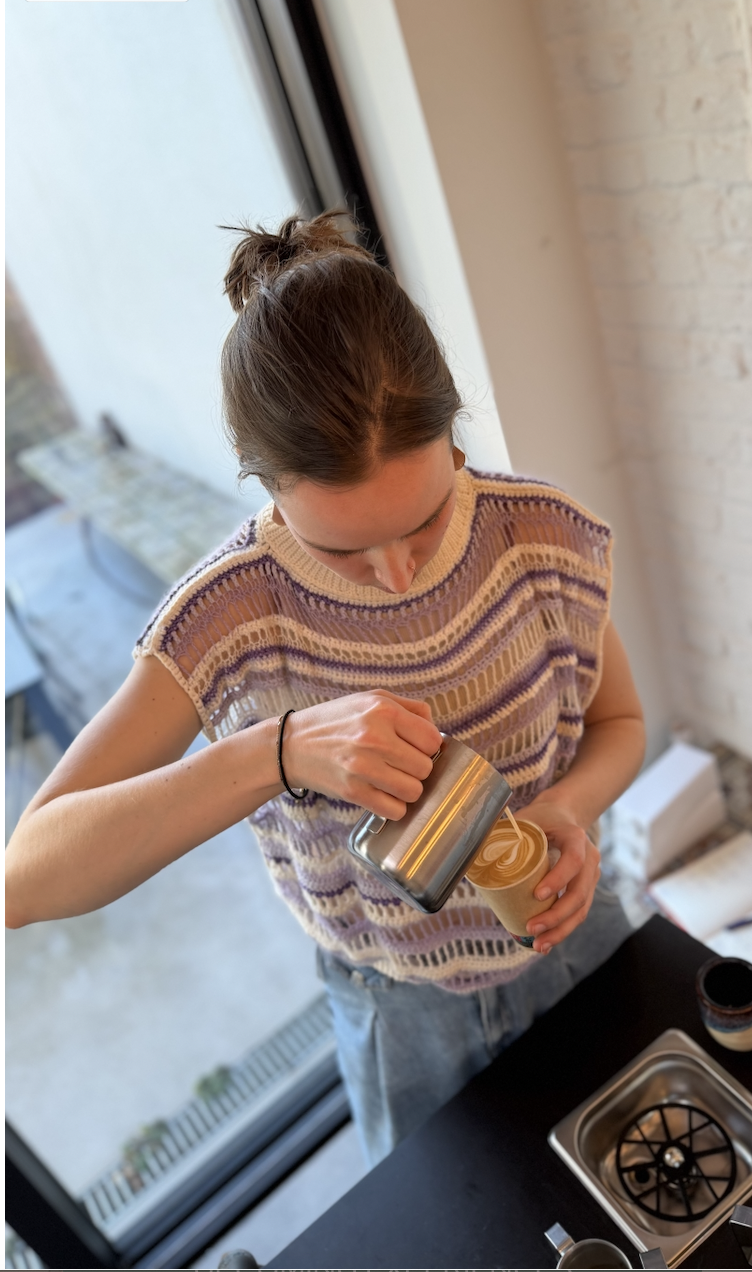 A barista is pouring steamed plant-based milk into a paper cup of coffee to make a latte art design. She stands near a large window in a kitchen.