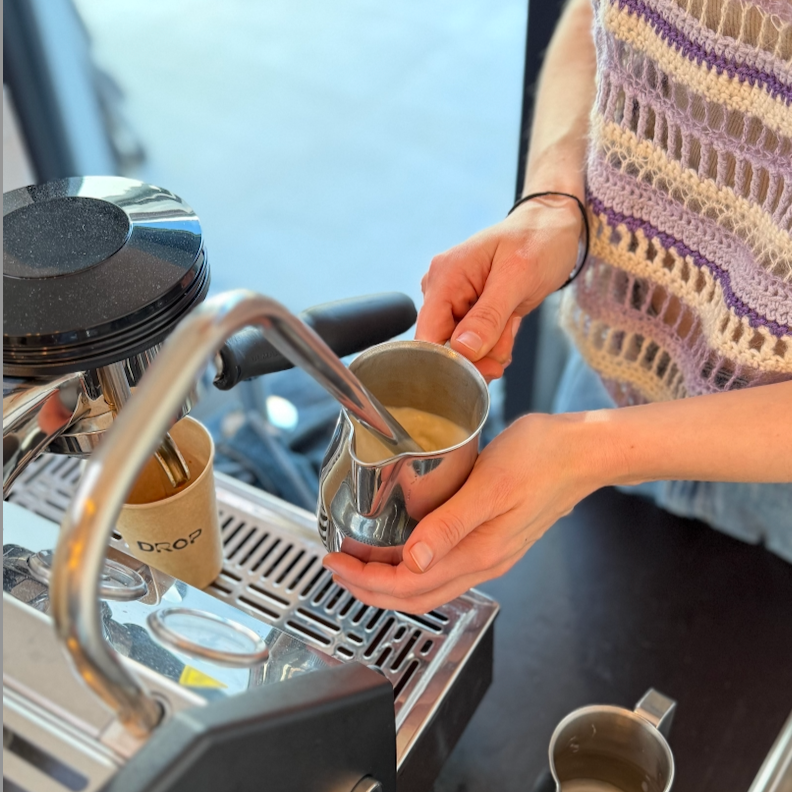 A barista making a specialty coffee drink. She is steaming plant-based milk, while the espresso is running into a paper coffee cup.