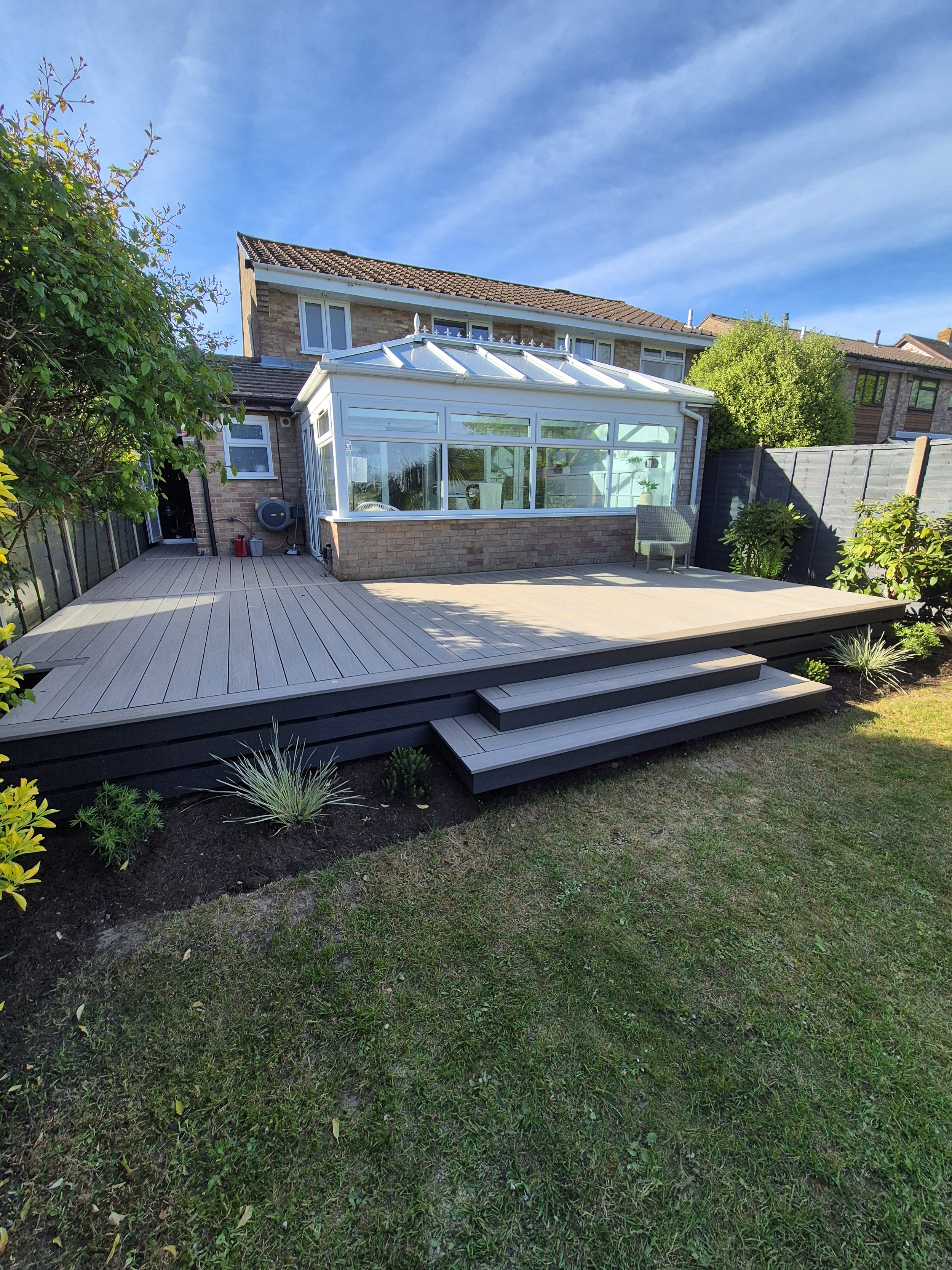 A modern backyard patio with white tiled flooring, a small grassy area, a wooden fence, and a covered seating area with outdoor furniture, surrounded by trees and neighboring houses.