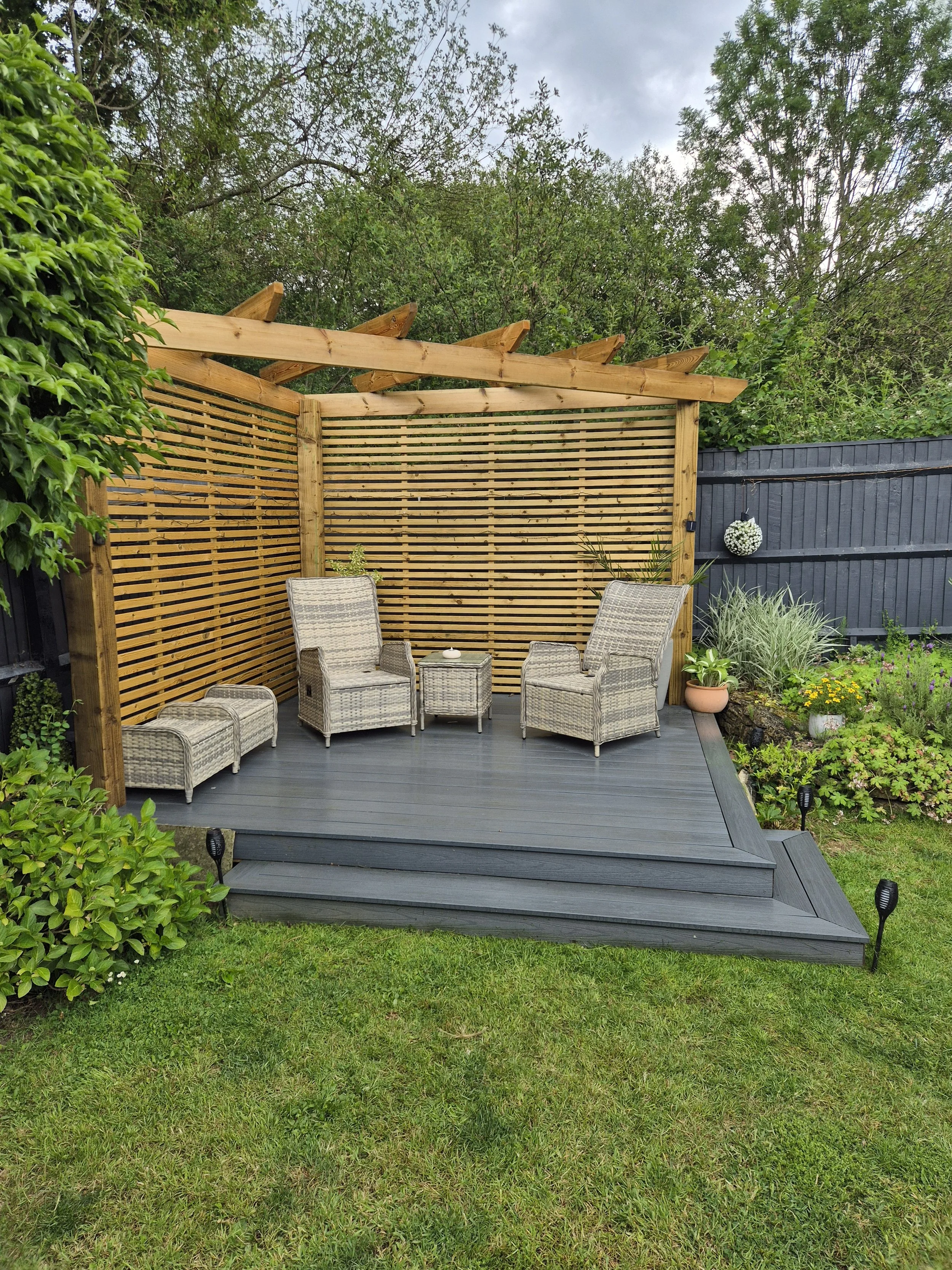 A modern outdoor patio with white tiled flooring, gray outdoor furniture including a sofa and ottomans, and a wooden slat fence surrounding the space with some small trees and potted plants.