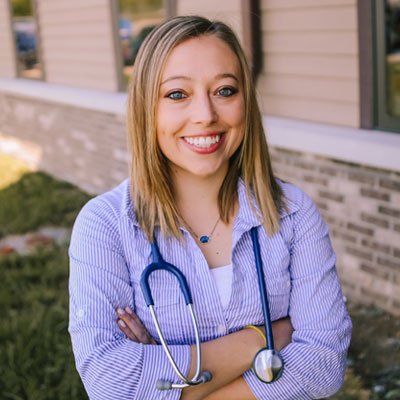 A young woman with blonde hair smiling outdoors, wearing a purple striped shirt with a stethoscope around her neck.