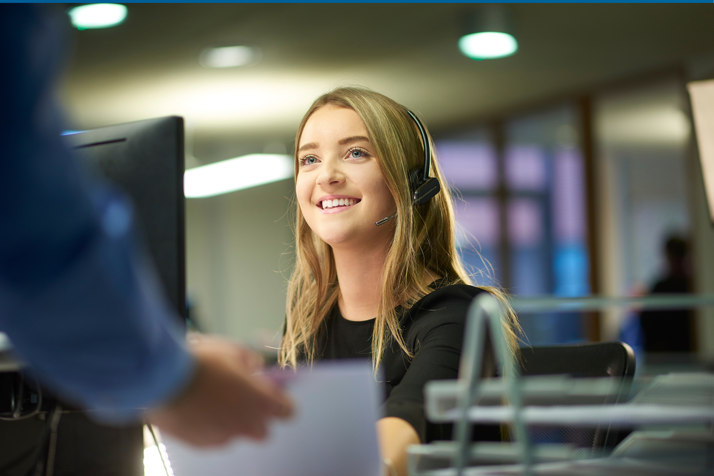 A young woman with long blonde hair wearing a headset, smiling and working at a computer in an office environment.