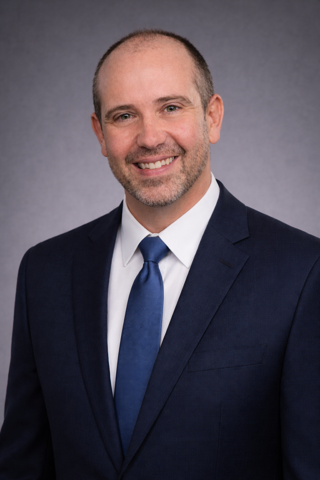 A professional headshot of a middle-aged man with short hair, a beard, and a friendly smile, wearing a navy suit, white shirt, and blue tie, against a gray background.