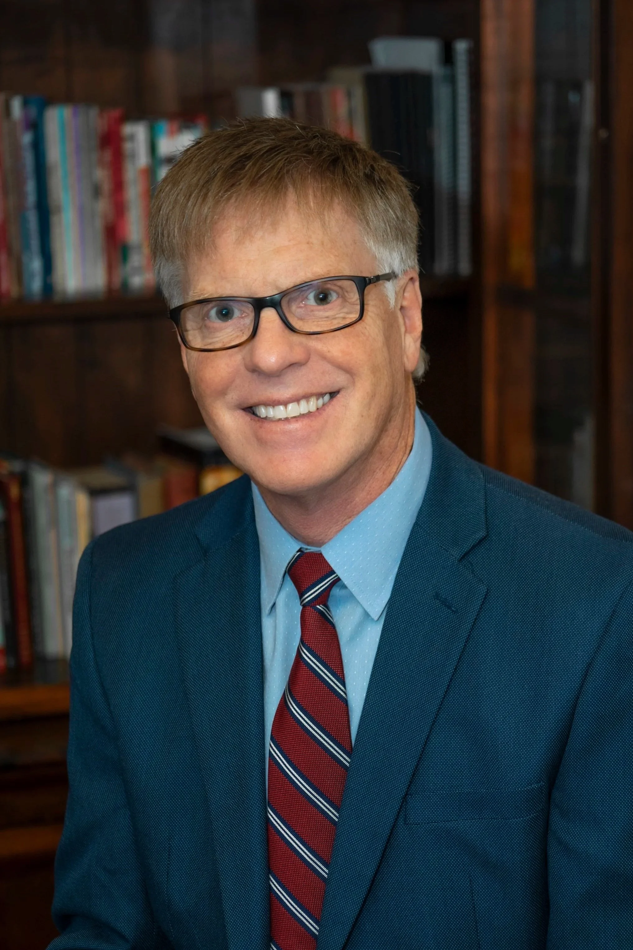 A smiling man with glasses and curly hair wearing a blue suit, light blue shirt, and a red, white, and blue striped tie. He is standing in front of a bookshelf filled with books.
