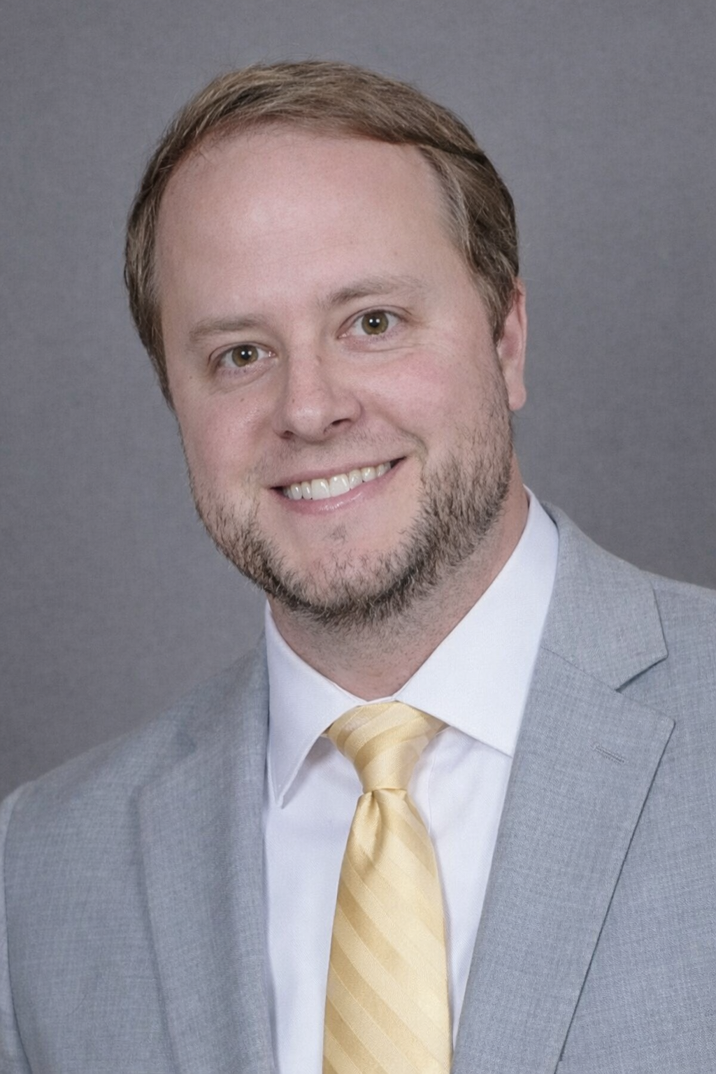 A man with light brown hair, a beard, and brown eyes, dressed in a light gray suit, white shirt, and yellow tie, smiling at the camera against a gray background.