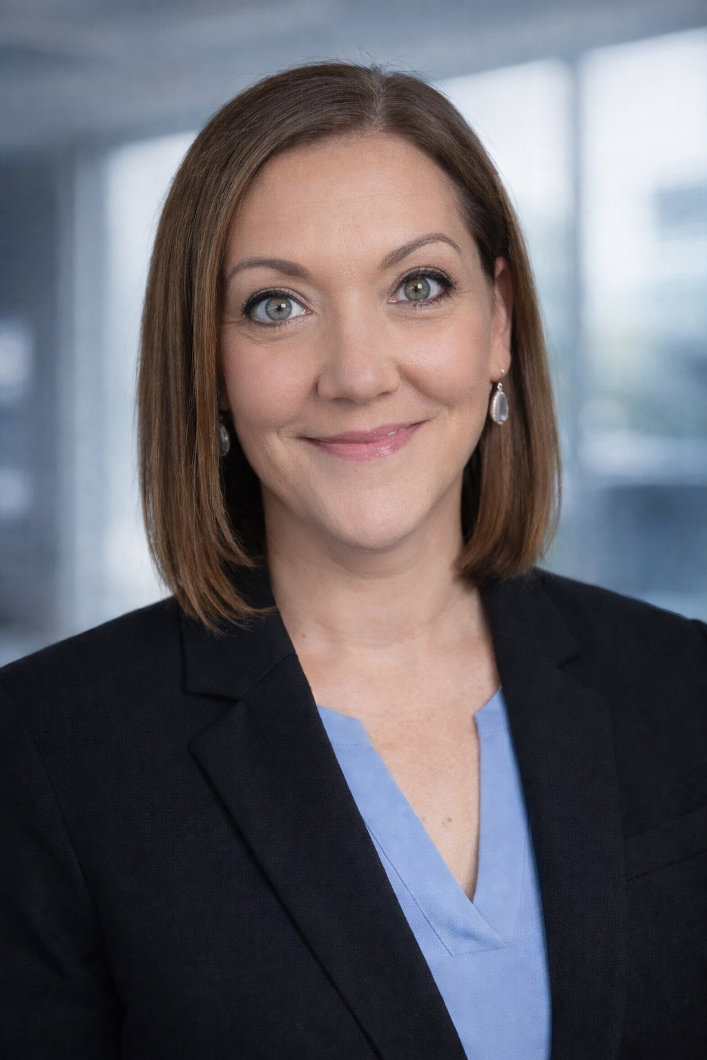 A professional woman with shoulder-length brown hair, wearing a black blazer and light blue blouse, smiling in front of a blurred modern office background.