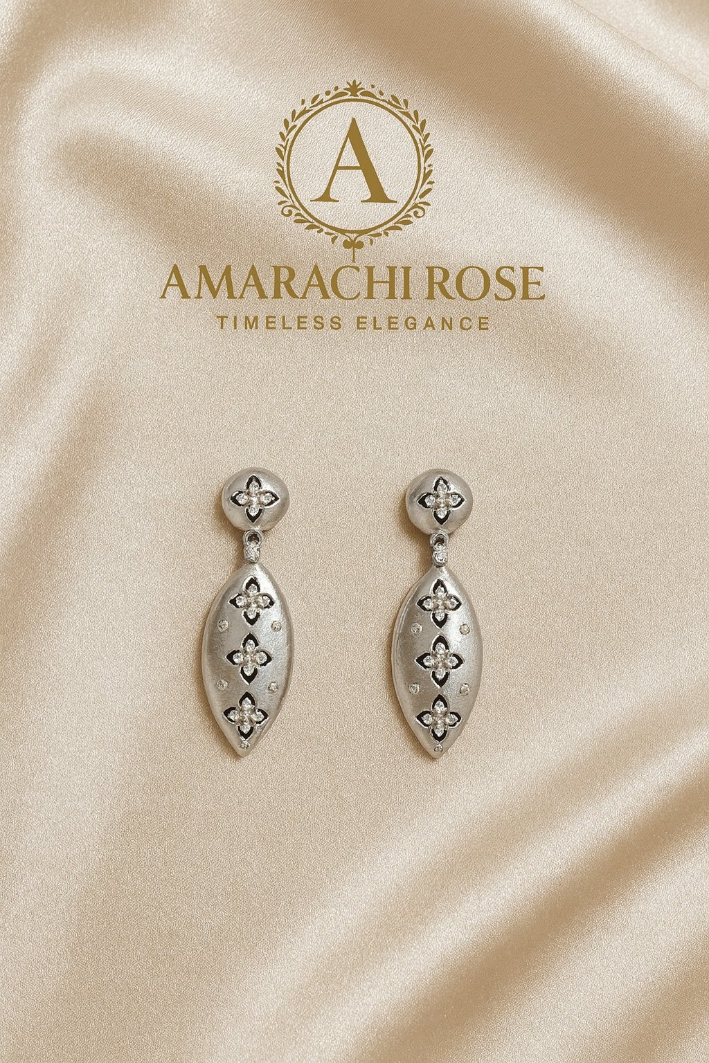 A pair of silver earrings with floral design, displayed on a satin beige background. The background features a logo with the letter A inside a decorative emblem, and the text 'AMARACH ROSE TIMELESS ELEGANCE'.