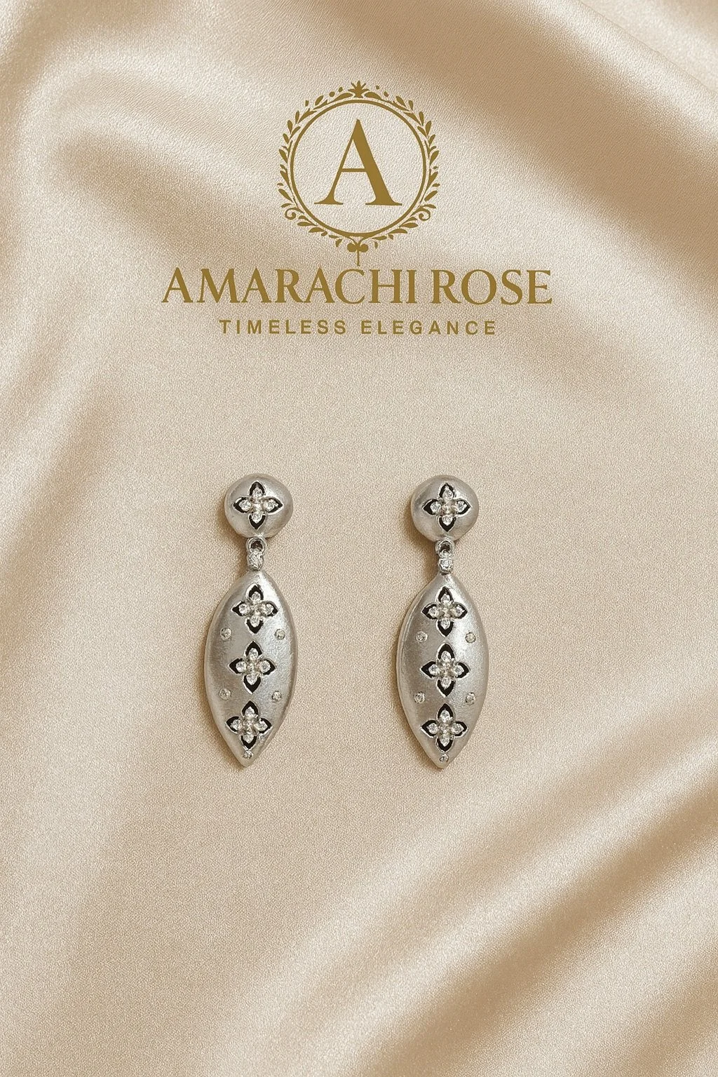 Silver drop earrings with floral cutout design on a satin background. The earrings are on a beige card with gold embossed text reading "Amarachi Rose", and a logo with a letter "A" inside a laurel wreath. The tagline "Timeless Elegance" is also on the card.