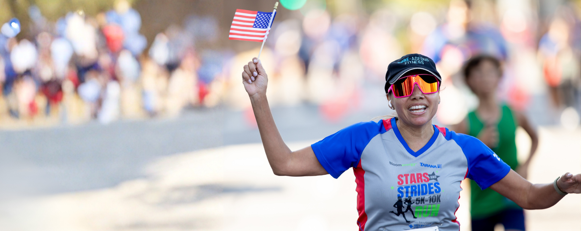 A woman smiling and celebrating as she runs, wearing sunglasses and a Stars & Strides shirt.
