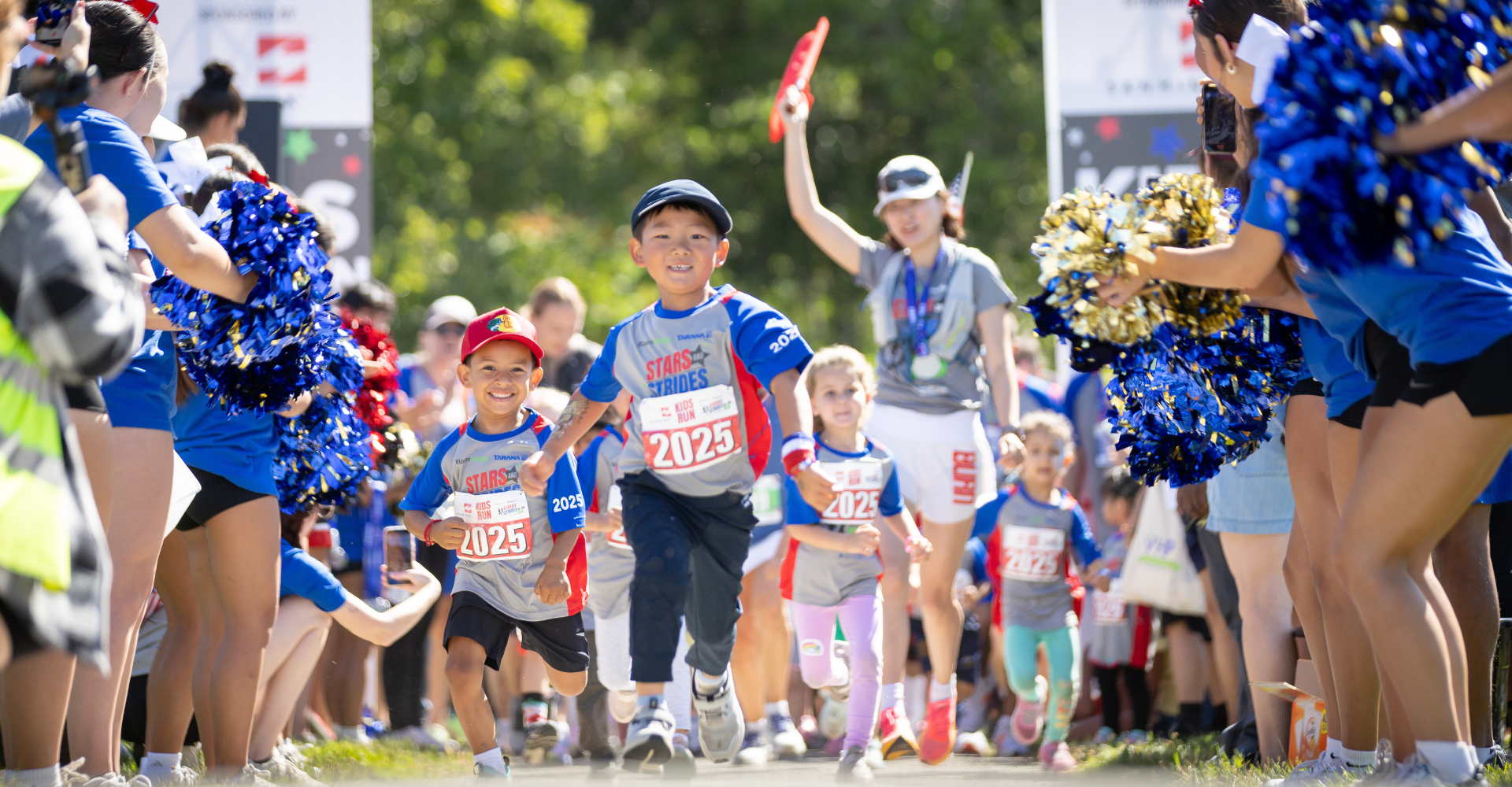 Children running in a race with cheerleaders on both sides clapping and holding pom-poms, in an outdoor setting on a sunny day.