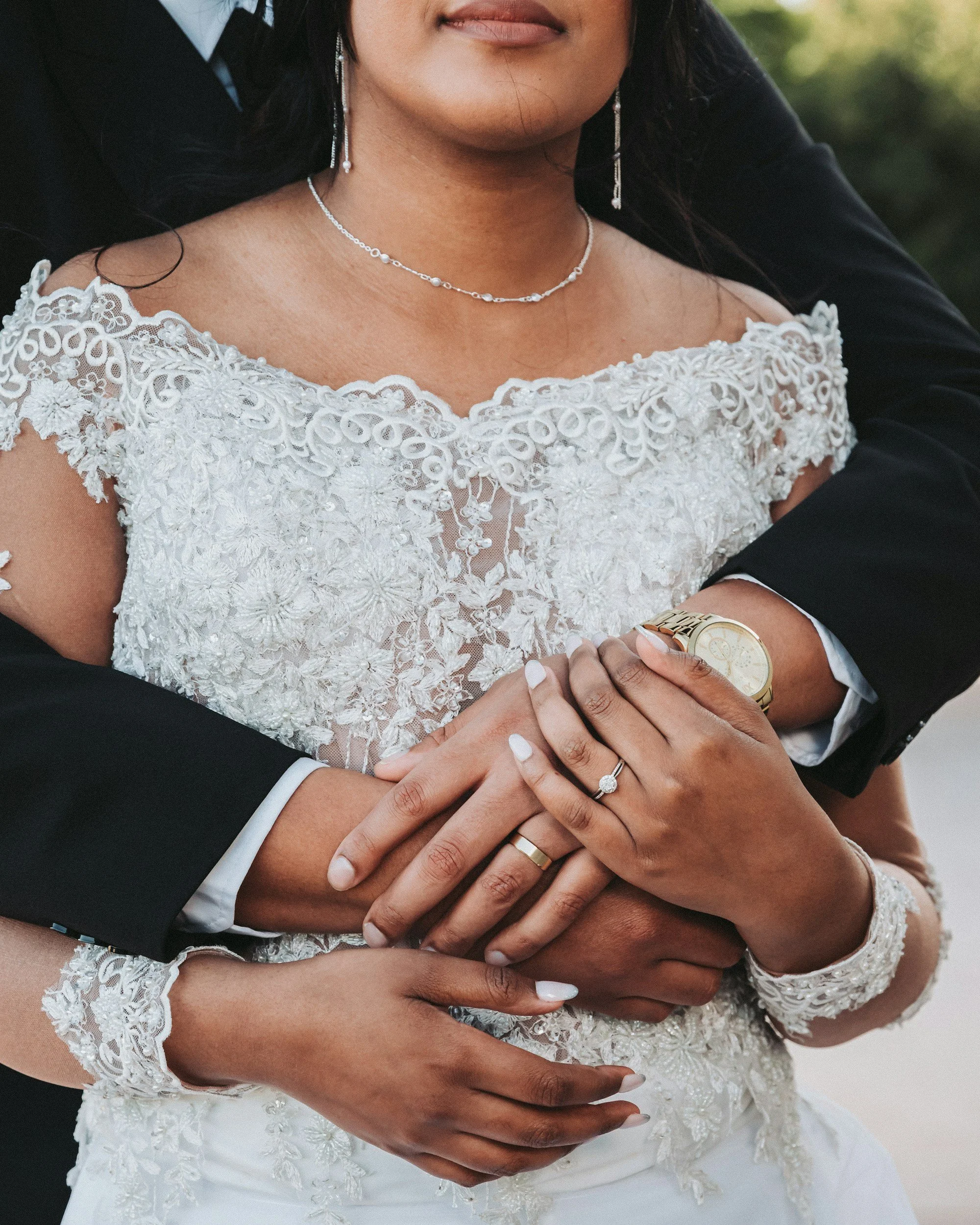 Close-up of a woman in a wedding dress and a man in a suit holding hands, showing wedding rings. The woman wears lace jewelry and a necklace, the man has a gold watch.