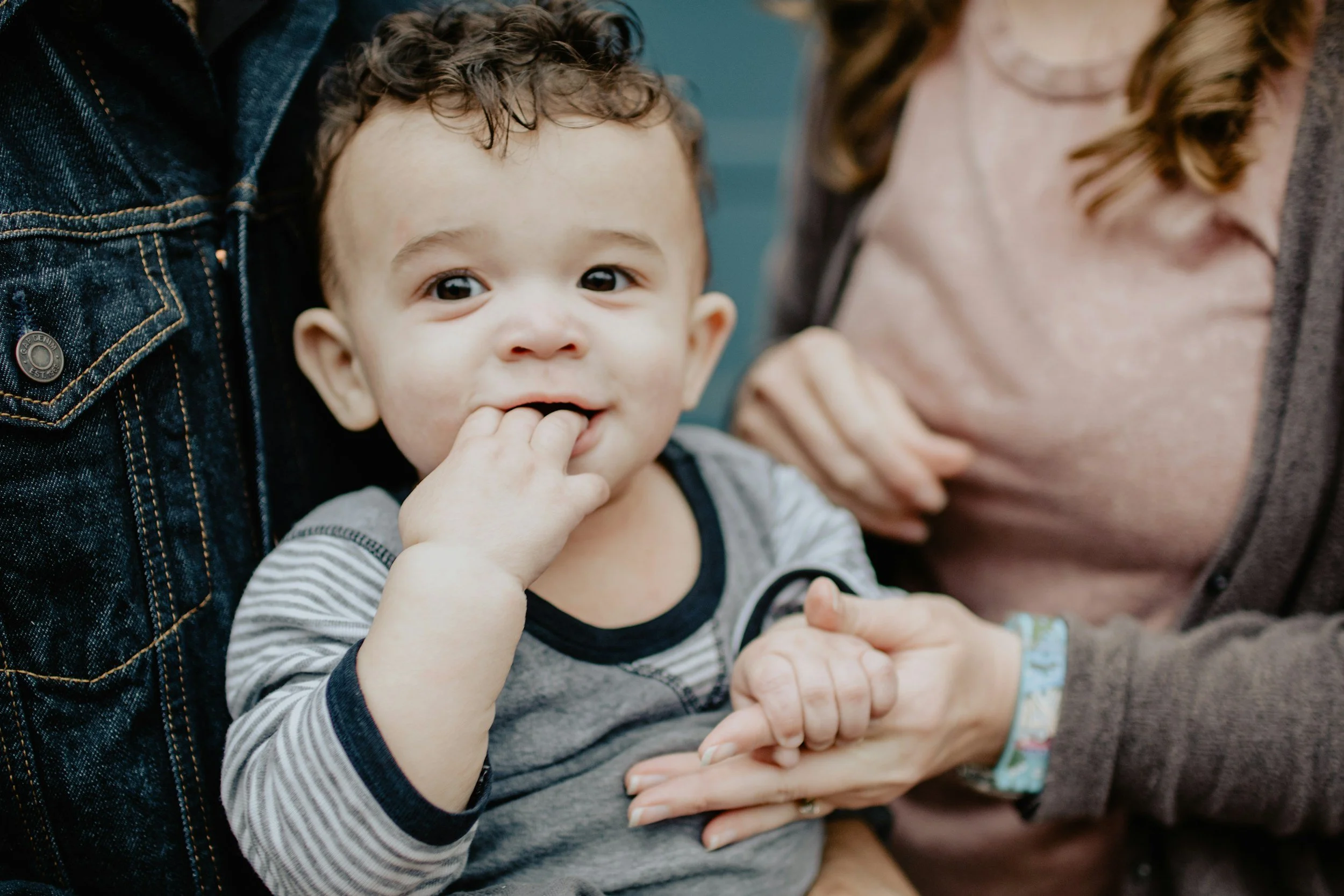 A young boy with curly hair and a striped shirt is smiling and has fingers in his mouth, surrounded by two adults. One adult is wearing a denim jacket, and the other is dressed in a brown sweater. The grown-ups appear to be holding and supporting the